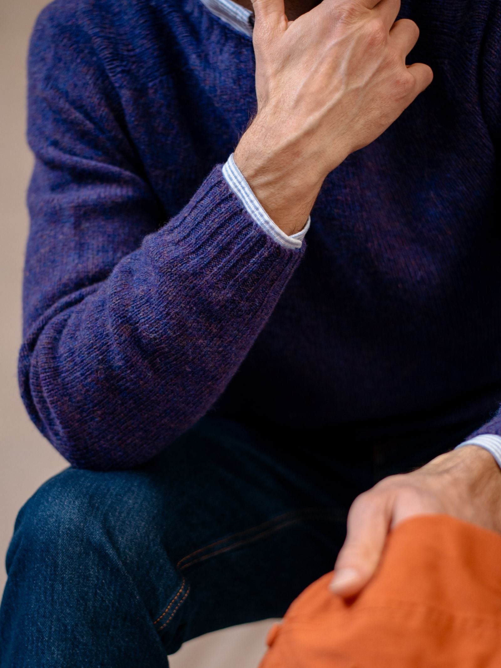 Wearing a blue Shetland Jumper by Campbells of Beauly, blue jeans, and a light blue collared shirt, a person—face partially out of frame—rests a hand on their chin and another on an orange item, highlighting heritage-inspired colours.