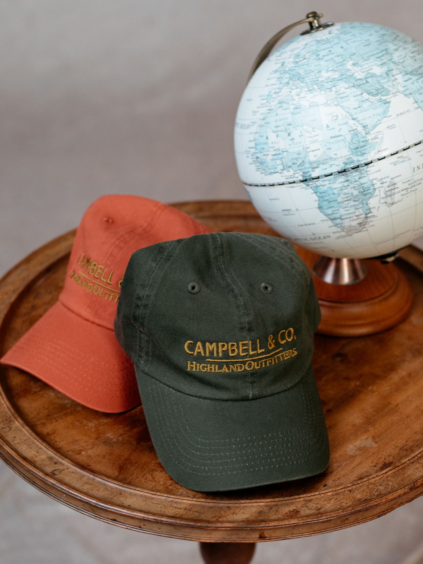 Two Campbells of Beauly baseball caps, one green and one orange, sit on a round wooden table next to a globe showing Africa and Europe—stylish and comfortable Campbells Baseball Caps for everyday wear.
