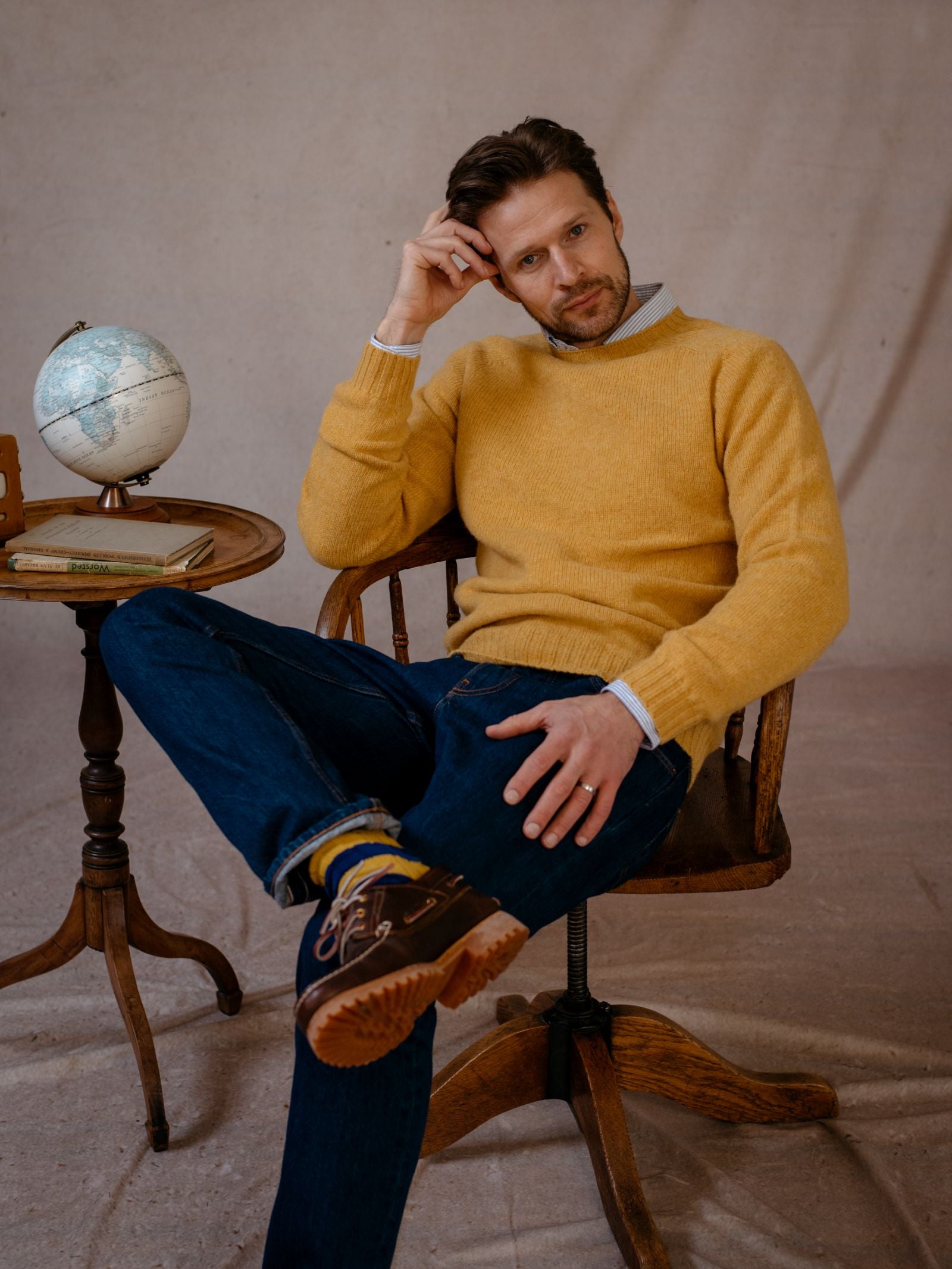A man in a Campbells of Beauly Shetland Jumper and jeans sits thoughtfully on a wooden chair, head resting on his hand. Next to him is a small table with a globe and books, all set against a plain backdrop.