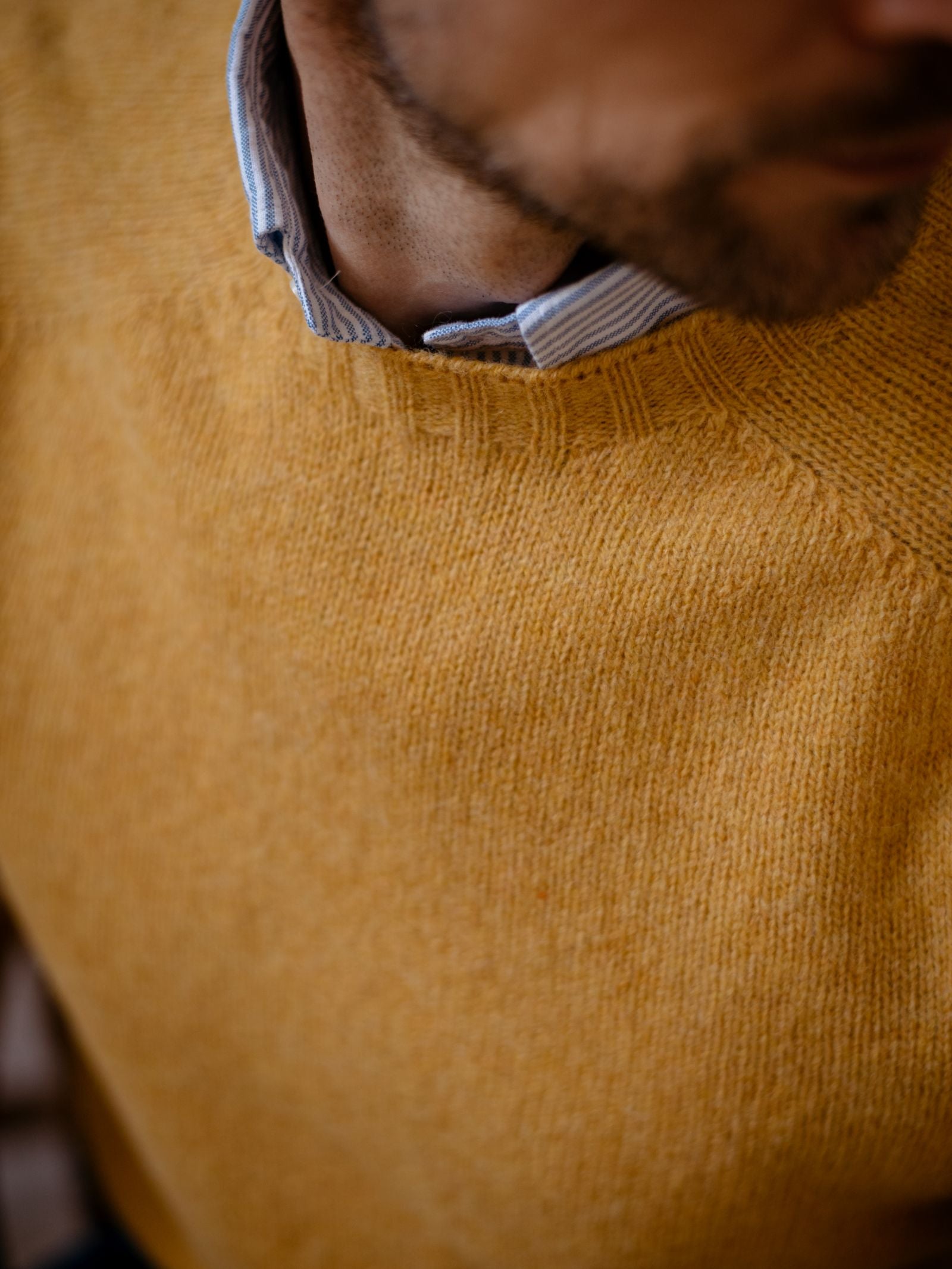 A close-up shows a person in the Campbells of Beauly Shetland Jumper, mustard yellow, layered over a light blue collared shirt. Only the lower face and neck with a trimmed beard are visible; eyes remain out of frame.