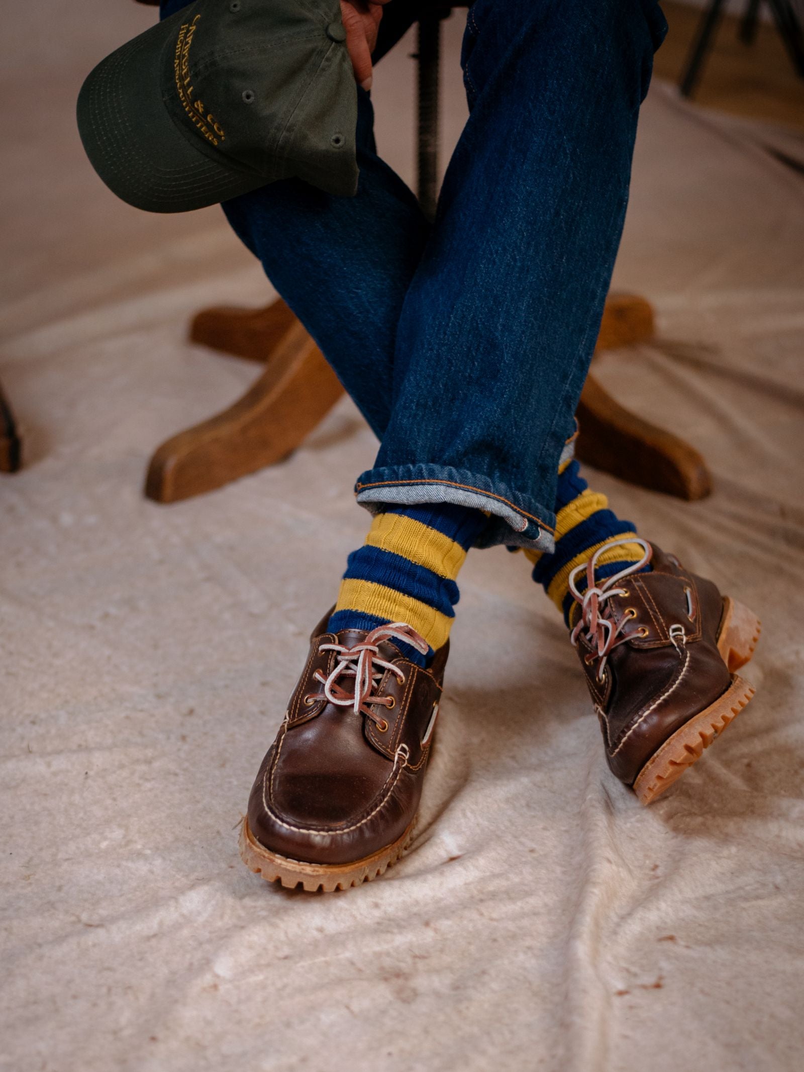 A person sits cross-legged on a wooden chair, wearing Campbells of Beauly Stripe Socks with reinforced heel and toe, blue jeans, brown leather shoes with light soles, and holding a green cap.