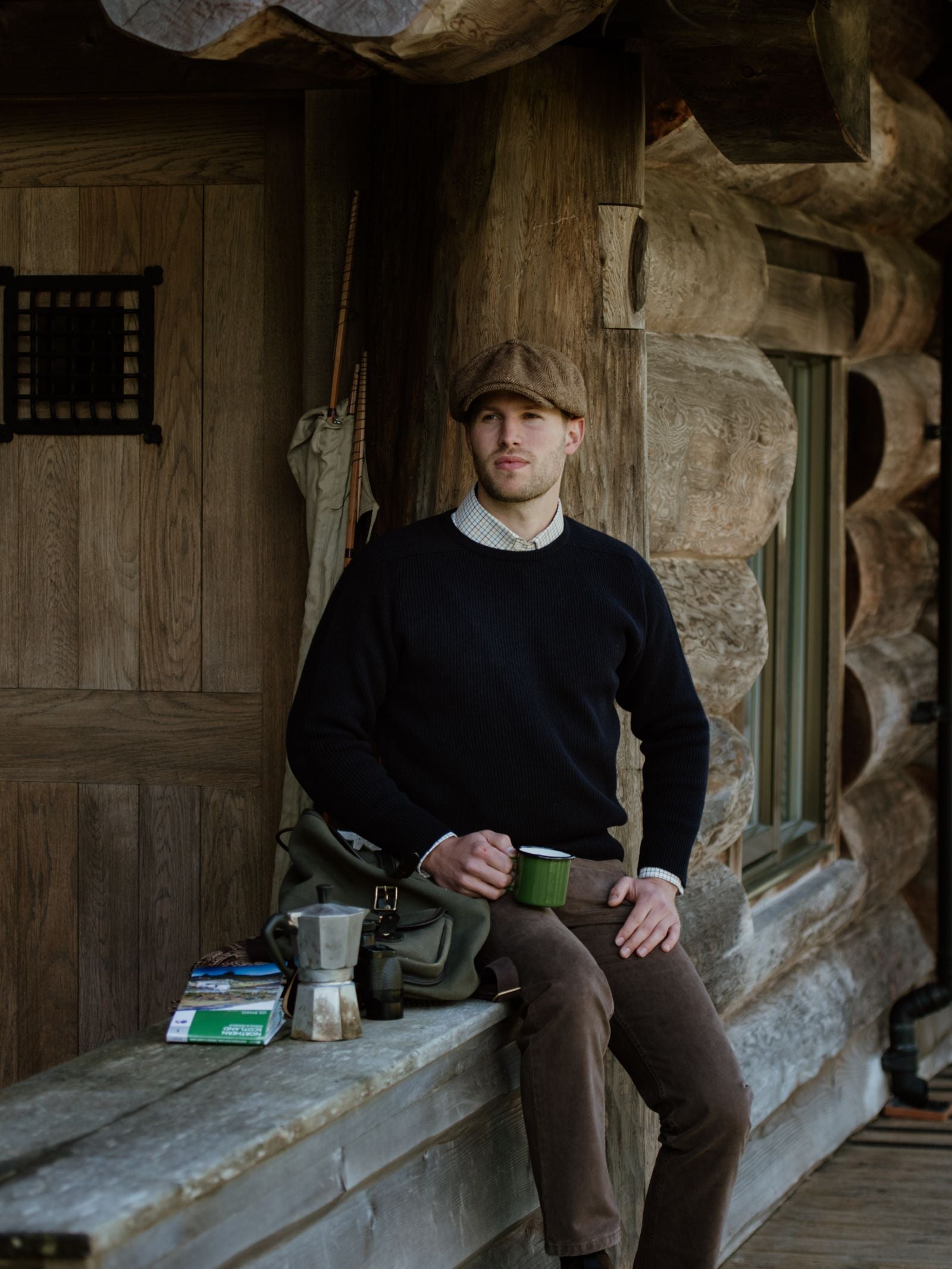 A man in a flat cap and dark sweater sits on a wooden porch, holding a green mug. He wears the Campbell’s of Beauly Fishermans Rib Crew Jumper, which suits the log cabin setting with books, a bag, and coffee maker nearby.
