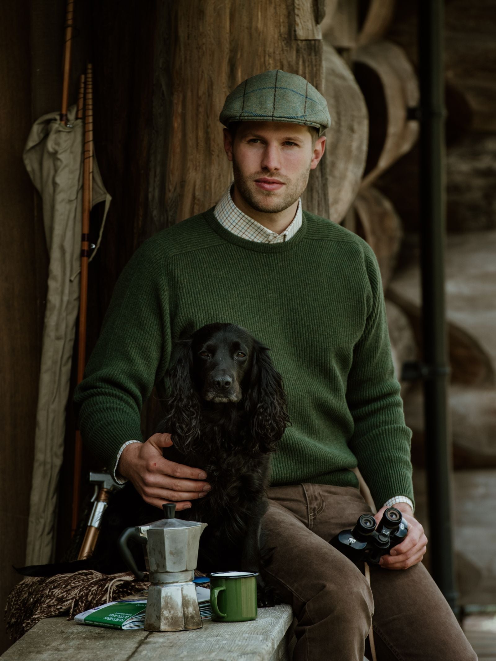 A man in a green Campbells of Beauly Fishermans Rib Crew Jumper with suede elbow patches sits on a rustic porch, holding binoculars and resting his hand on a black dog, with coffee and outdoor gear on the table beside him.
