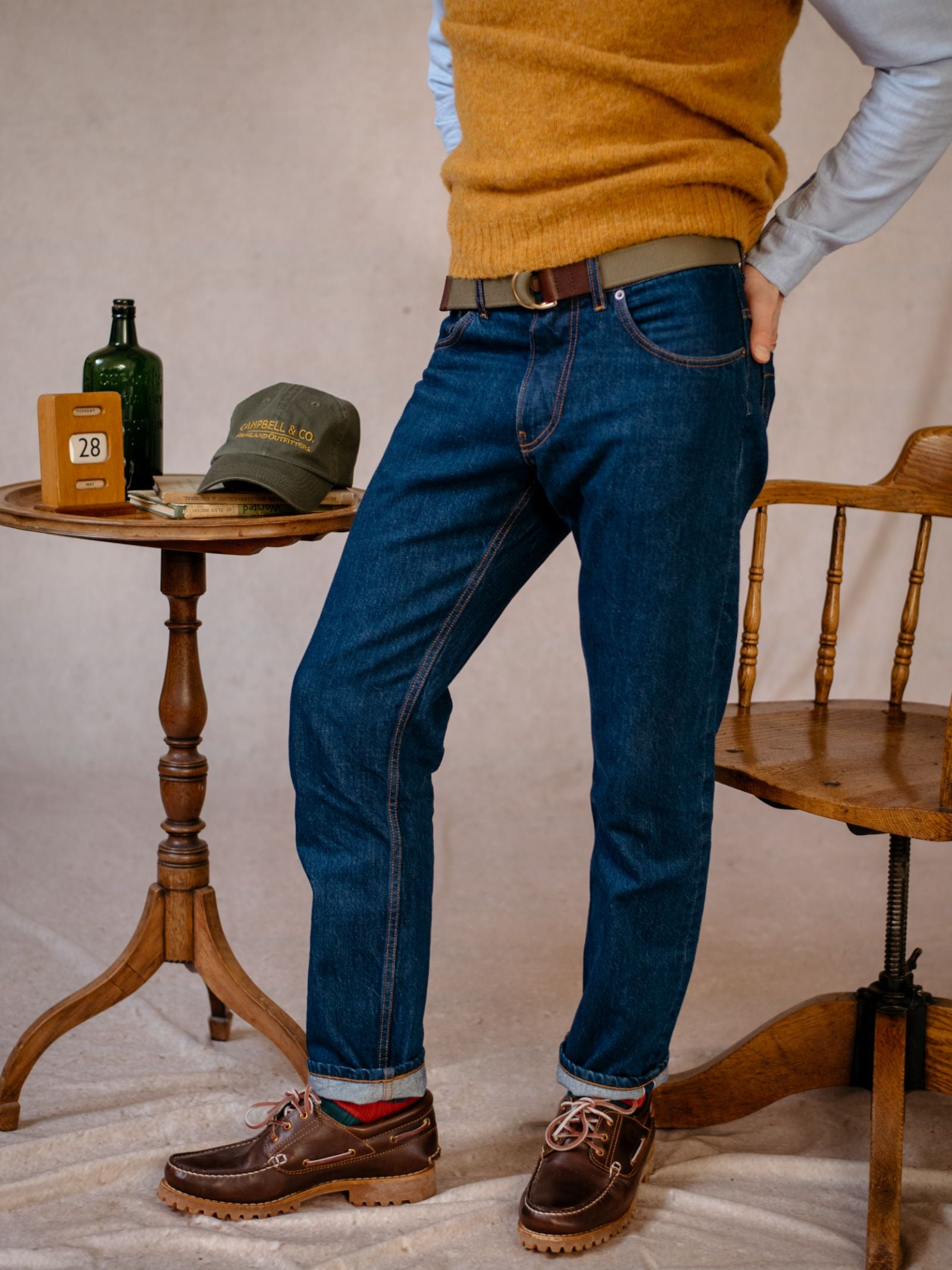 A person in a mustard sweater vest, Campbells of Beauly Denim Jeans, and brown shoes stands beside a small wooden table with a cap, books, bottle, and calendar. A wooden chair is nearby. The persons face is not visible.
