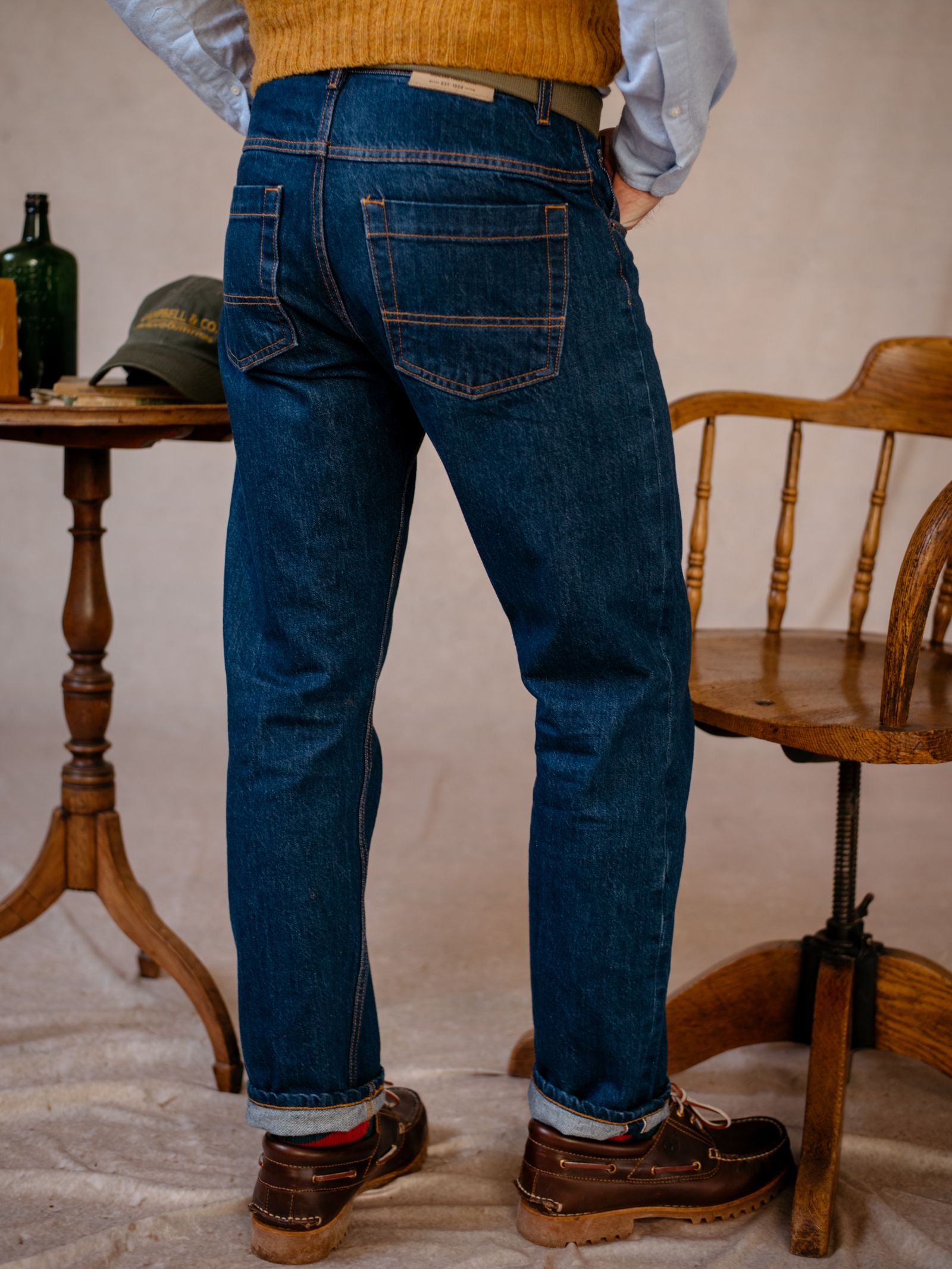 A person wearing Campbells of Beauly Denim Jeans, brown shoes, a light shirt, and a mustard sweater stands next to a wooden chair and a small round table with books and a bottle. The photo is taken from the back.