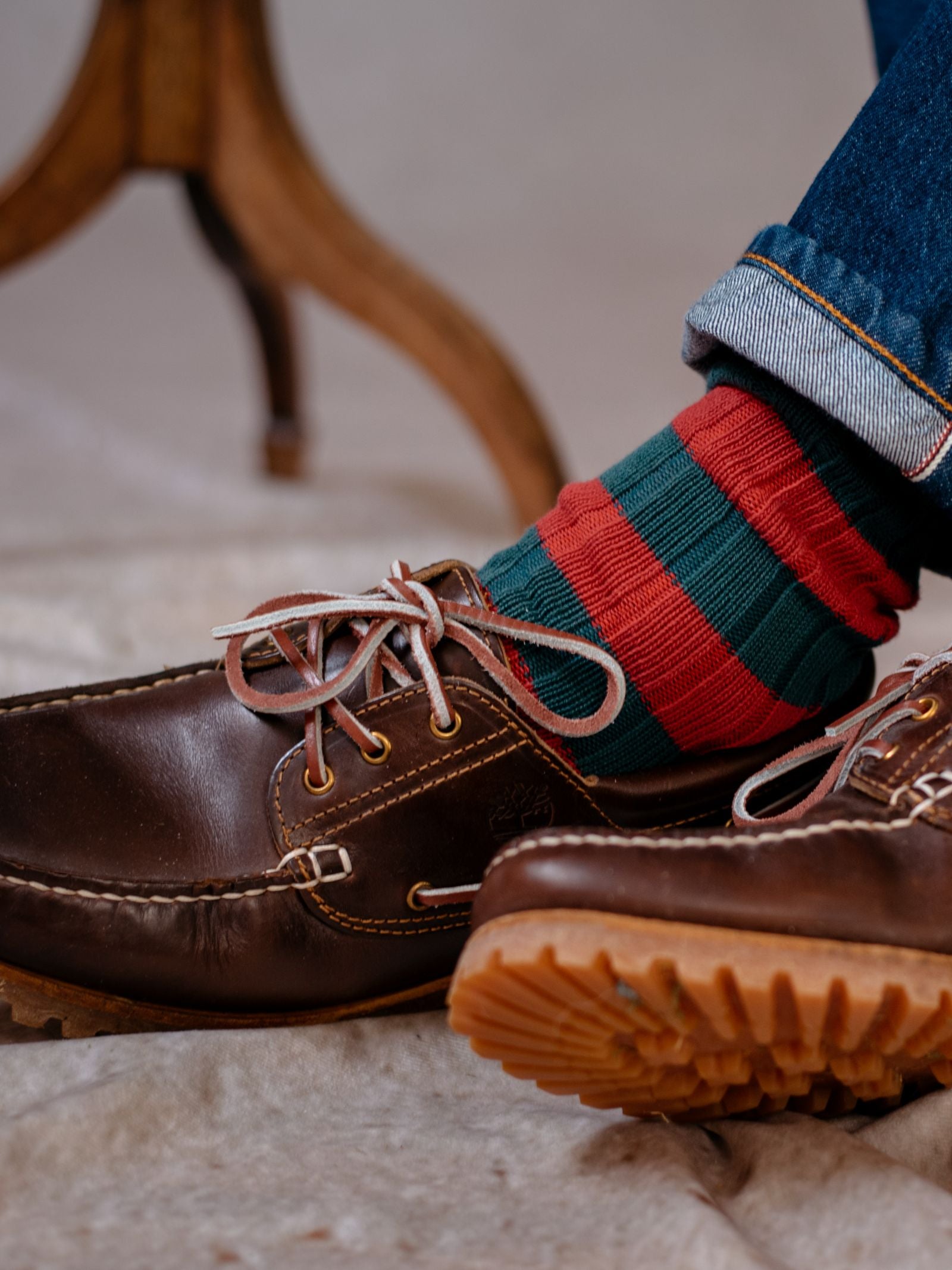 A close-up of a person wearing Campbells of Beauly Stripe Socks with reinforced heel, brown leather boat shoes, and blue rolled-up jeans, with part of a wooden table leg visible in the background.