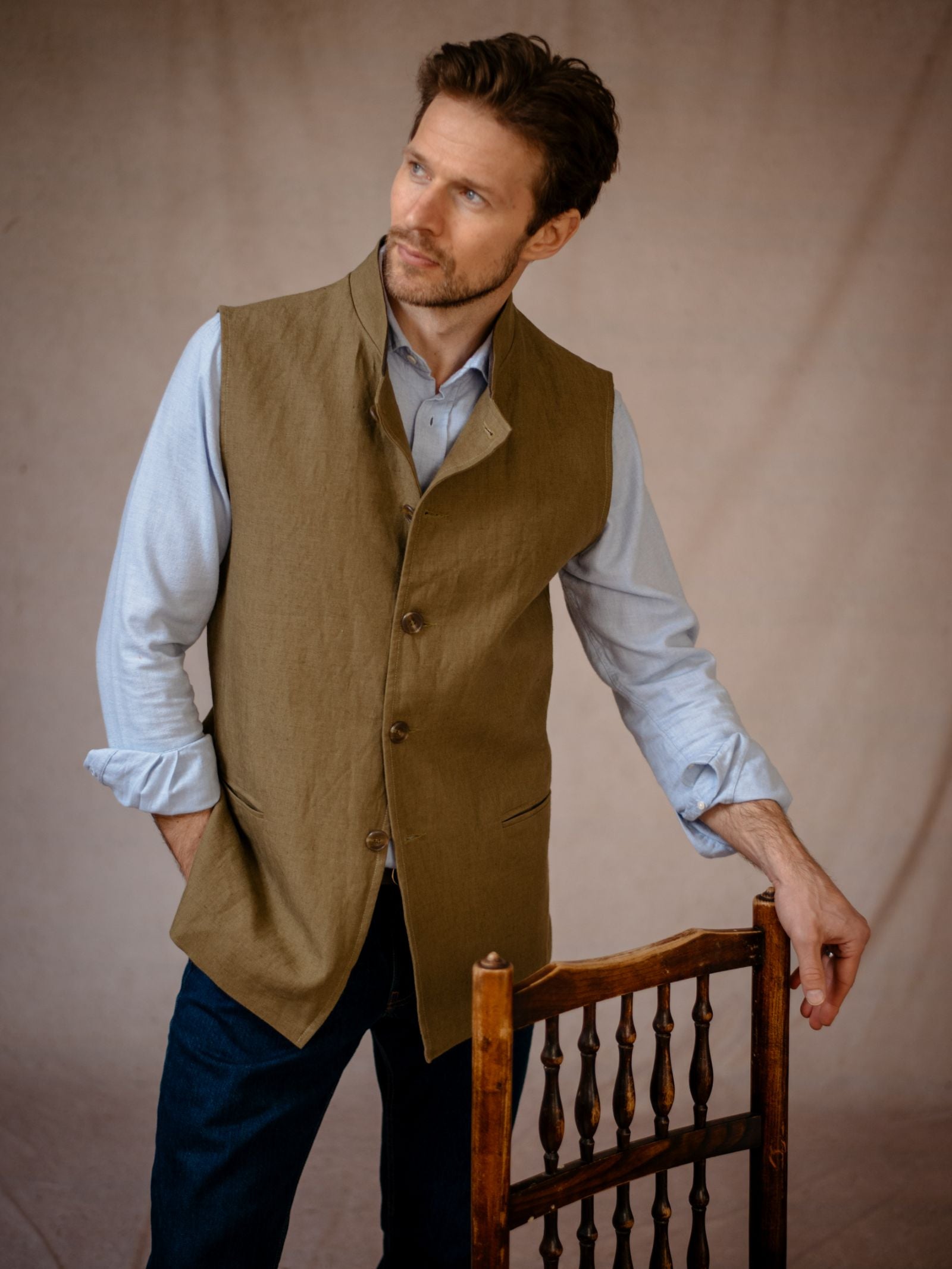 A man with light brown hair and a trimmed beard wears a light blue button-up shirt with rolled sleeves and the Linen Nehru Gilet from Campbells of Beauly, standing by a wooden chair and gazing thoughtfully to the side.