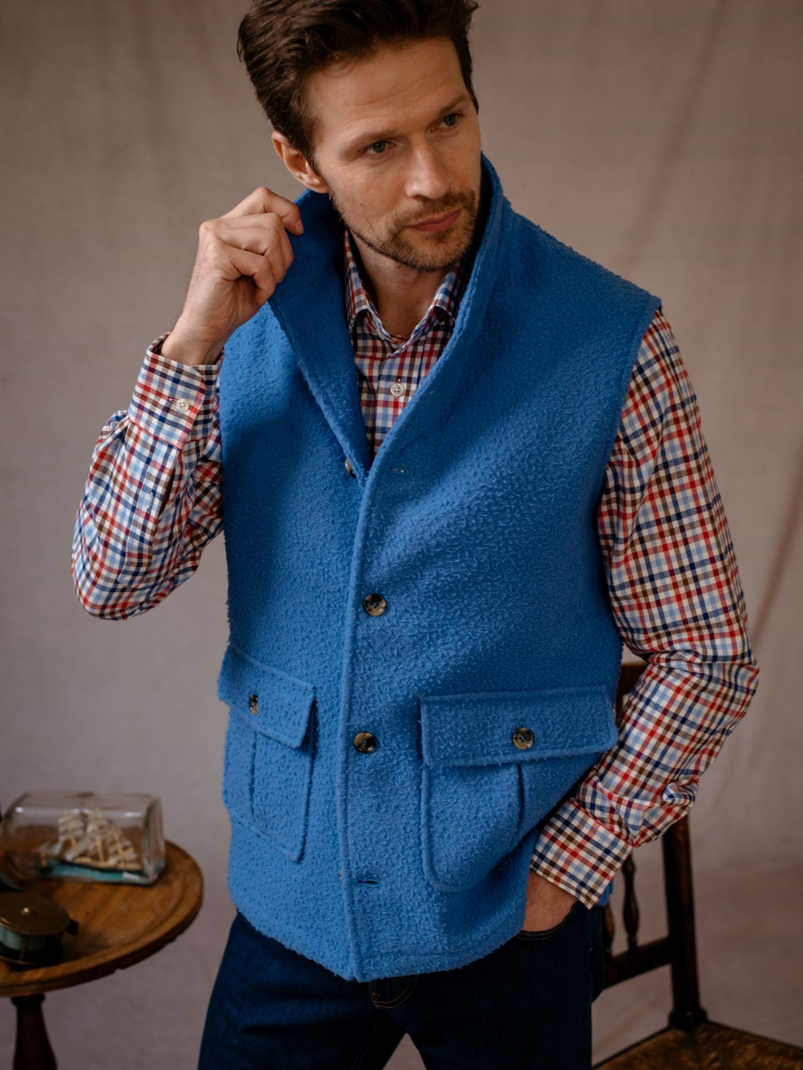A man with short brown hair and a trimmed beard stands indoors adjusting his collar, wearing a blue Casetino Fleece Gilet by Campbells of Beauly over a red and blue plaid shirt. A small table and chair are in the background.