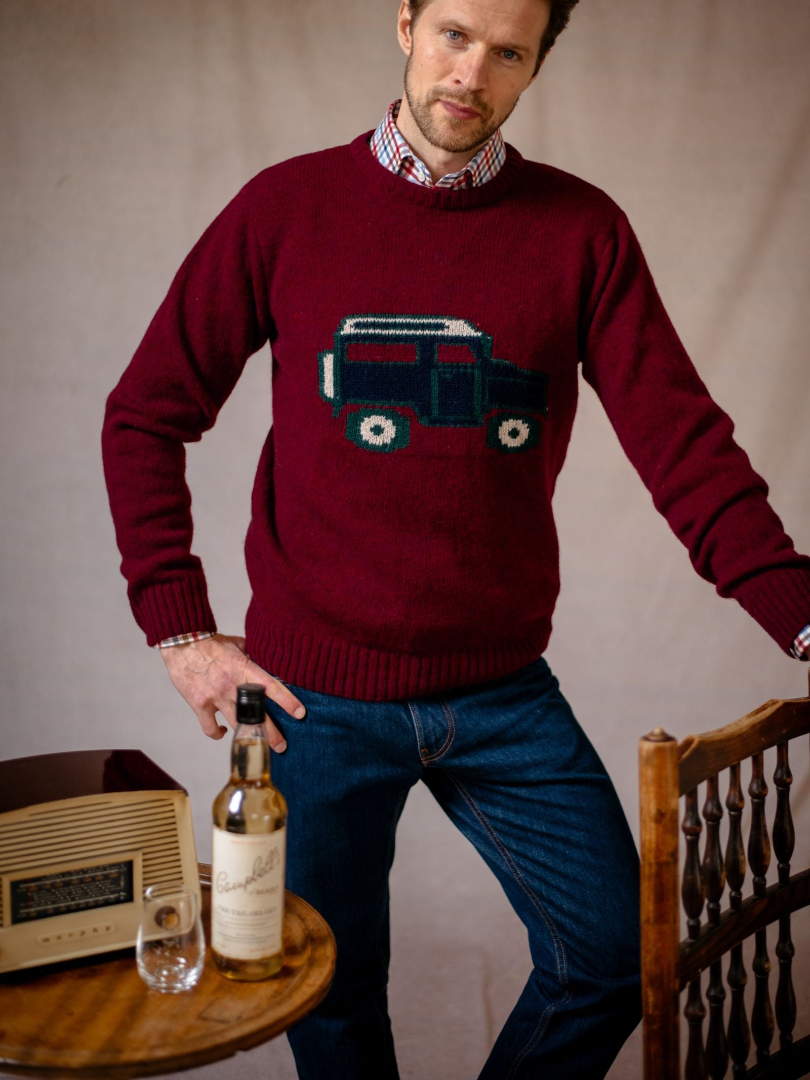 A man in the Campbells of Beauly Land Rover Defender Jumper, jeans, and a plaid shirt stands by a wooden chair. On the table are a vintage radio and whiskey—evoking classic British motoring style.