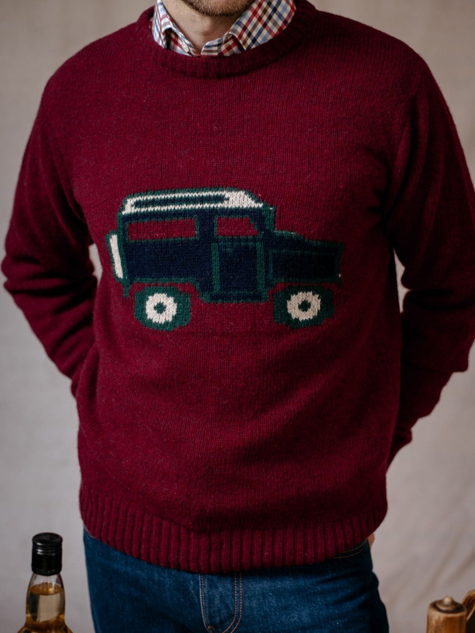 A person wears the Campbells of Beauly Land Rover Defender Jumper, which is burgundy with a blue and green Defender design, layered over a plaid shirt. Only their lower face and torso are seen; bottles are partially visible at the bottom.