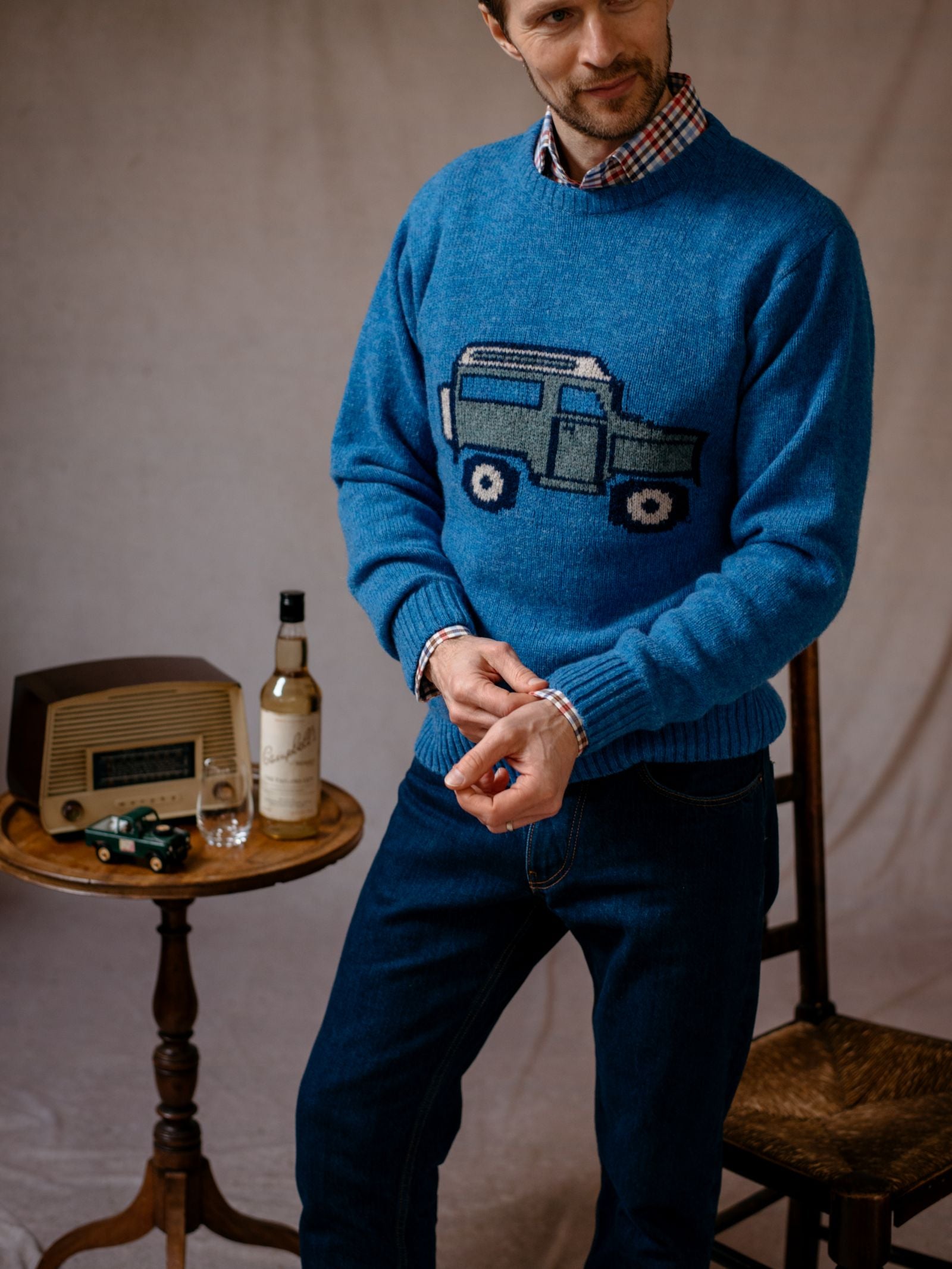 A man in a Campbells of Beauly Land Rover Defender Jumper stands by a vintage wooden table with a radio, whisky bottle, and toy cars, adjusting his cuff and smiling—a nod to classic British motoring style.
