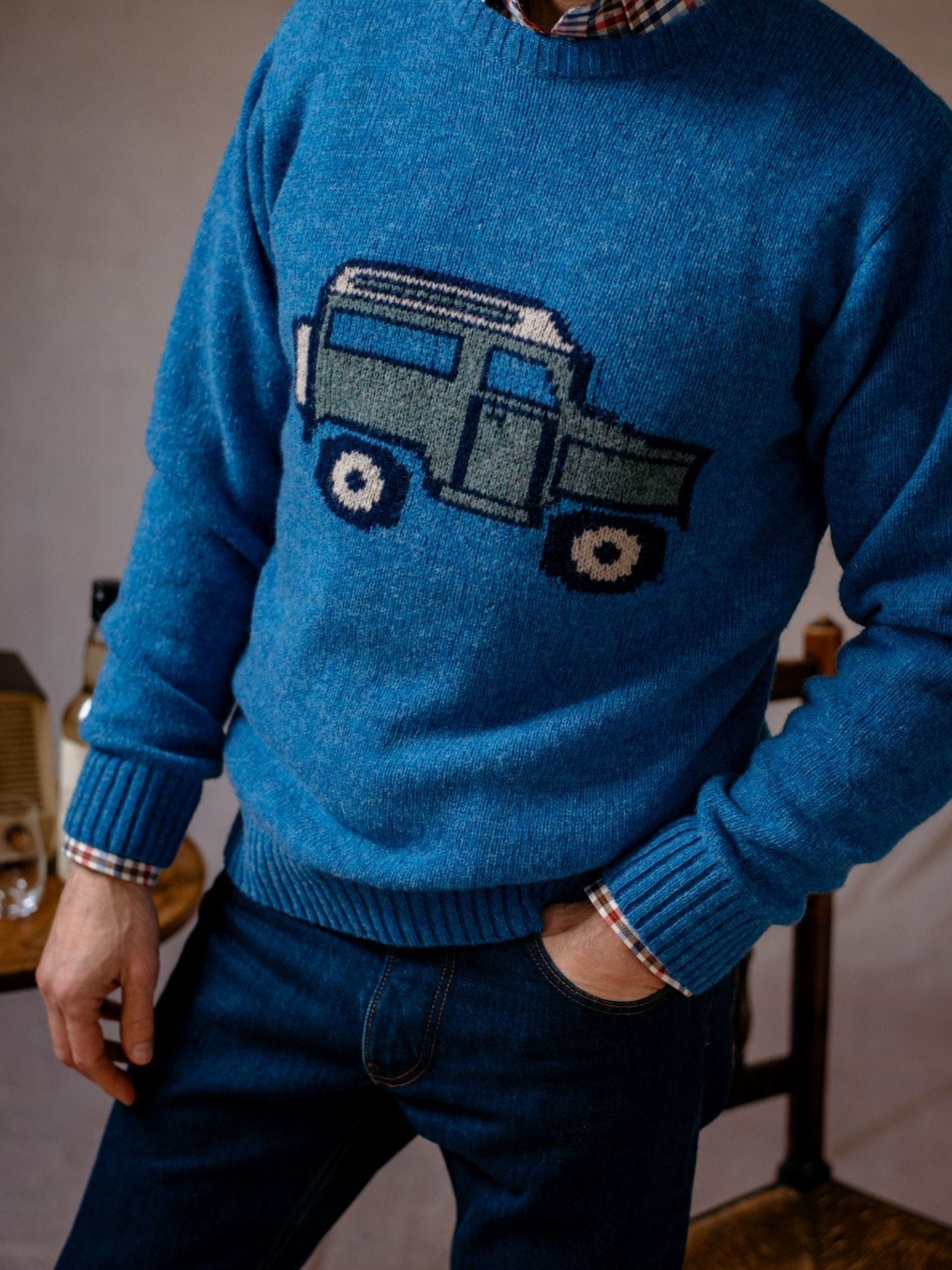 A person wears the Campbells of Beauly Land Rover Defender Jumper—a blue knit sweater with a Land Rover Defender graphic—layered over a plaid shirt and dark jeans, standing indoors by wooden furniture, evoking British motoring heritage.