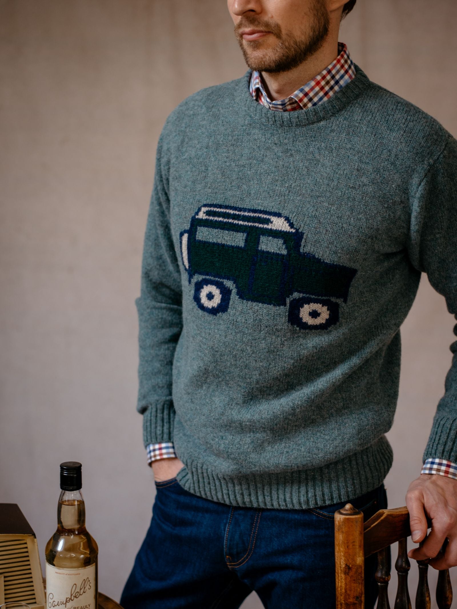 A man in a Campbells of Beauly Land Rover Defender Jumper—gray with a classic SUV graphic—layered over a plaid shirt and jeans stands by a table with a bottle of Scotch whisky. His face is partially out of frame.