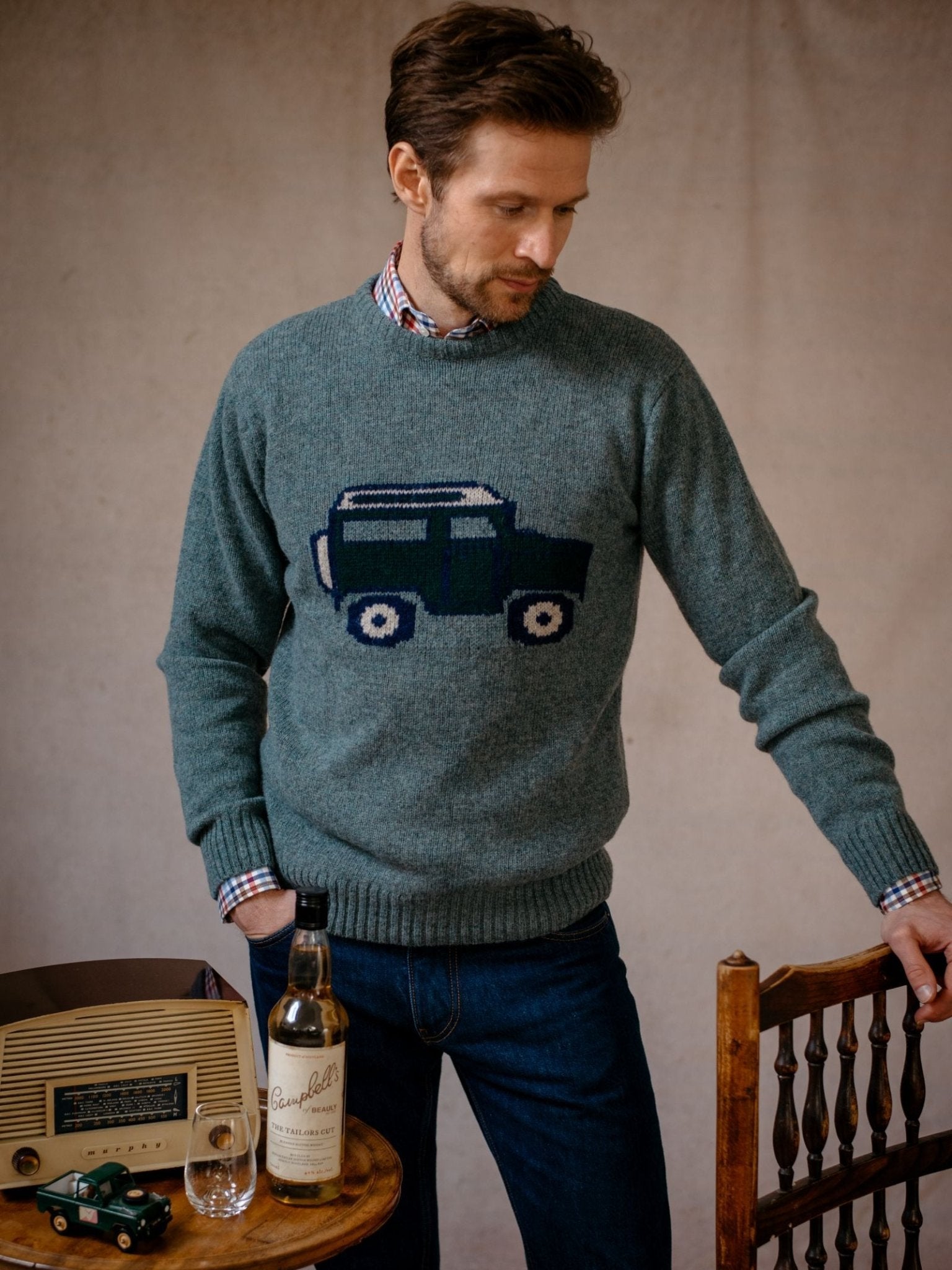 A man wearing the Land Rover Defender Jumper by Campbells of Beauly stands by a wooden chair, looking down. On the table are a vintage radio, green toy car, glass, and a bottle of Campbell’s whiskey.