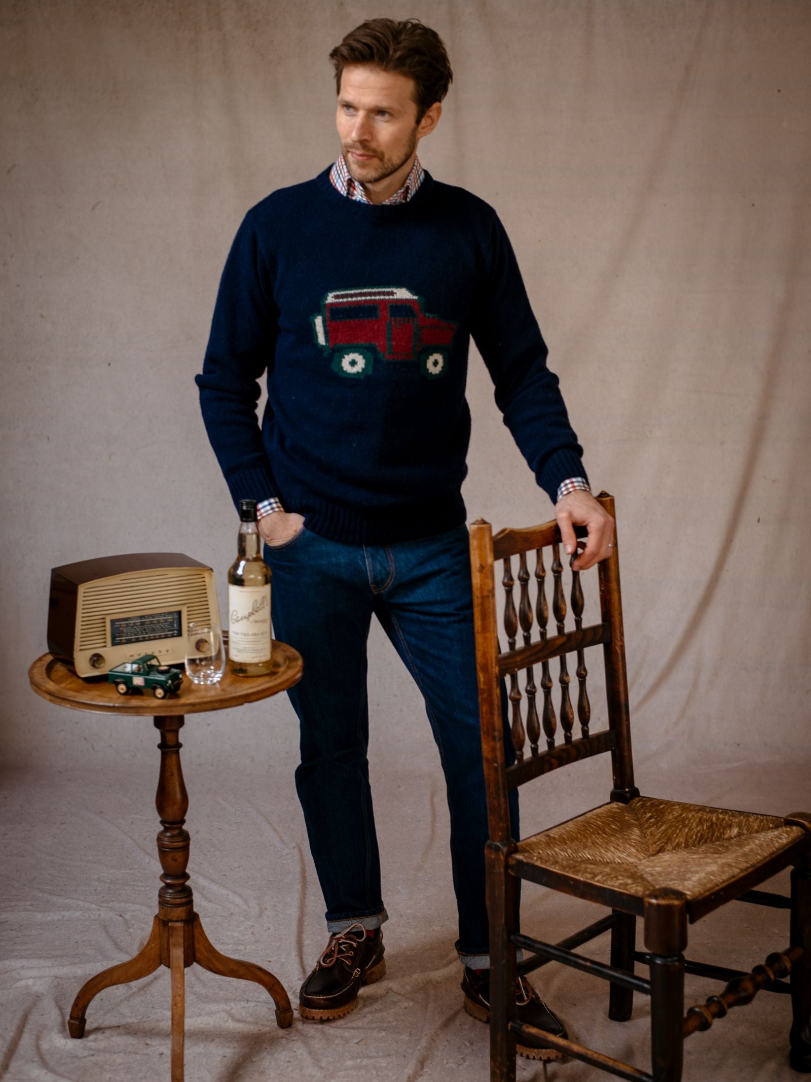 A man in a navy Campbells of Beauly Land Rover Defender Jumper, featuring a red and green vehicle, stands indoors by a wooden chair and round table with a vintage radio, bottle, glass, and British motoring-inspired toy car.