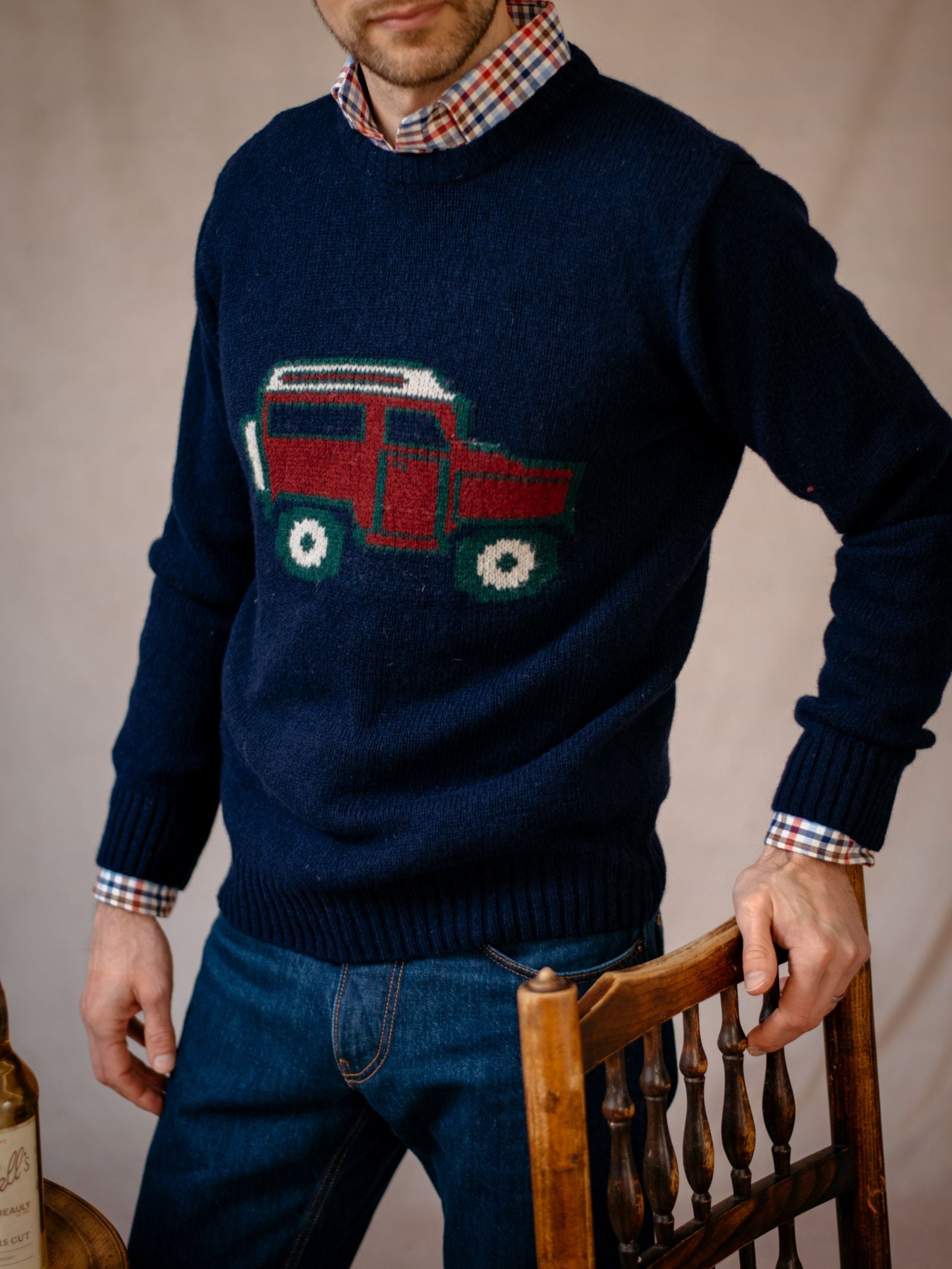 A man wearing the Campbells of Beauly Land Rover Defender Jumper, styled over a plaid shirt and blue jeans, stands by a wooden chair. His face is partially out of frame, highlighting classic British motoring style.