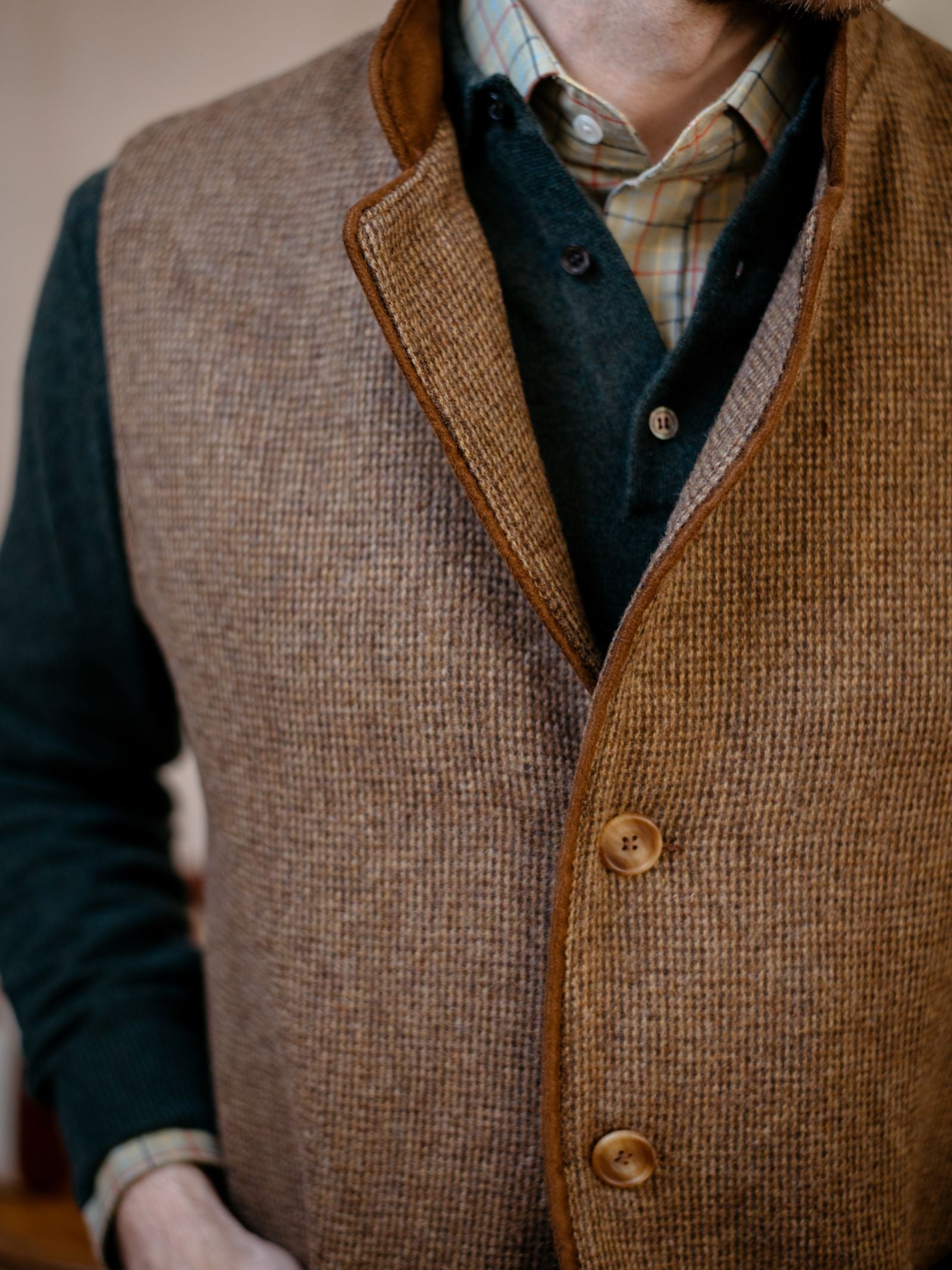 A man models the Campbells of Beauly Tweed Nehru Gilet over dark green and plaid collared shirts, one hand in his pocket. The image, cropped to show his torso and neck, highlights this versatile layering piece.