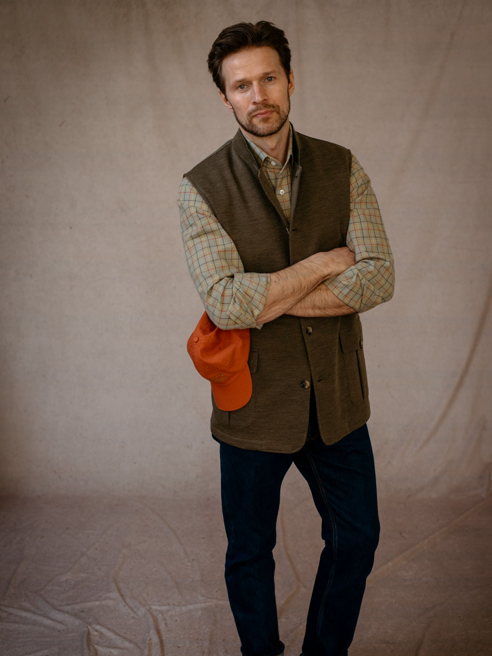 A man with short brown hair and a beard stands with arms crossed, wearing a plaid shirt, dark jeans, and the Campbells of Beauly Knitted Nehru Gilet as a stylish layering piece, holding an orange cap against a neutral backdrop.