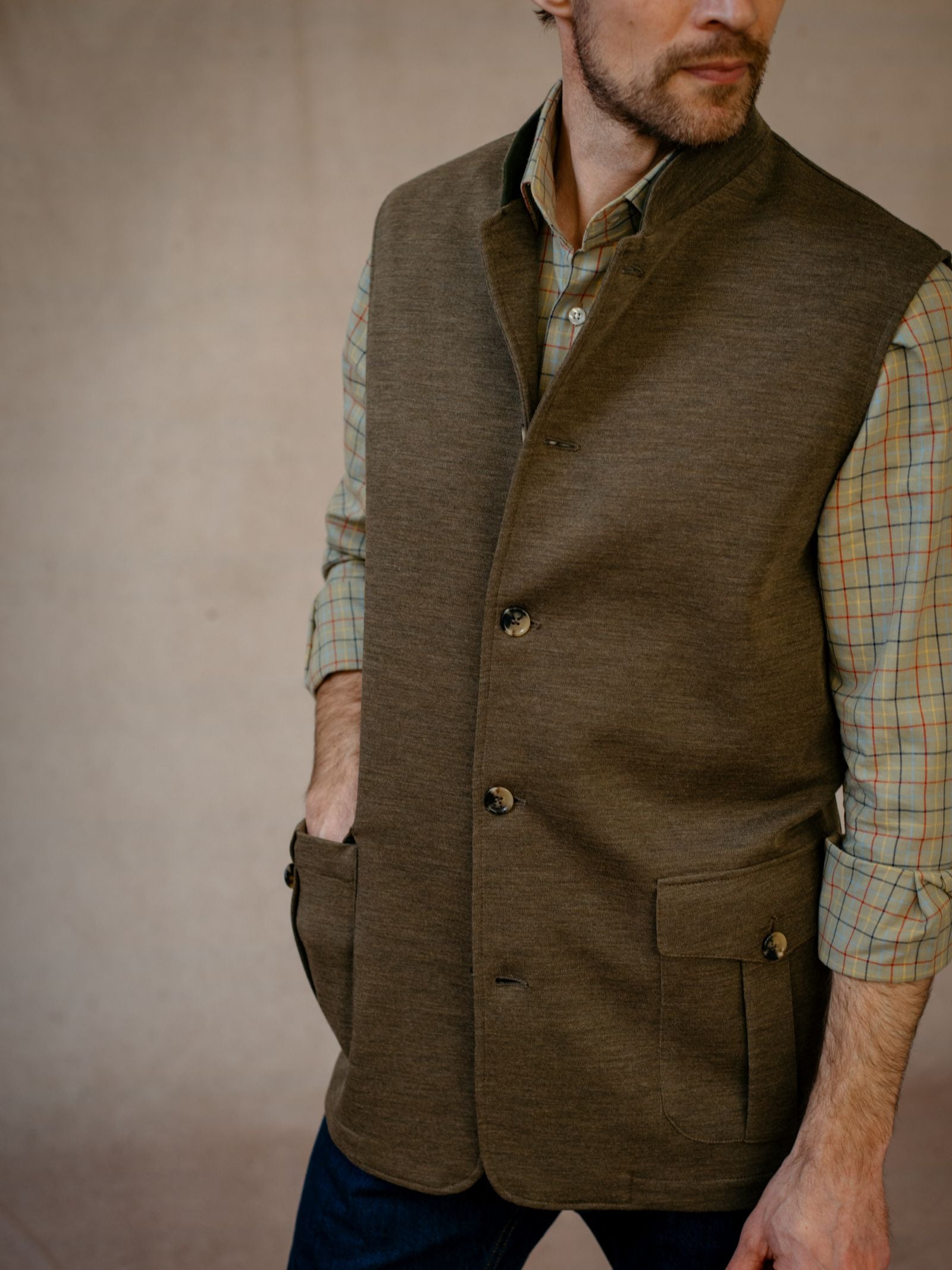 A man in a Campbell’s of Beauly Knitted Nehru Gilet over a green plaid shirt stands with one hand in his pocket, turned slightly away from the camera against a neutral background.