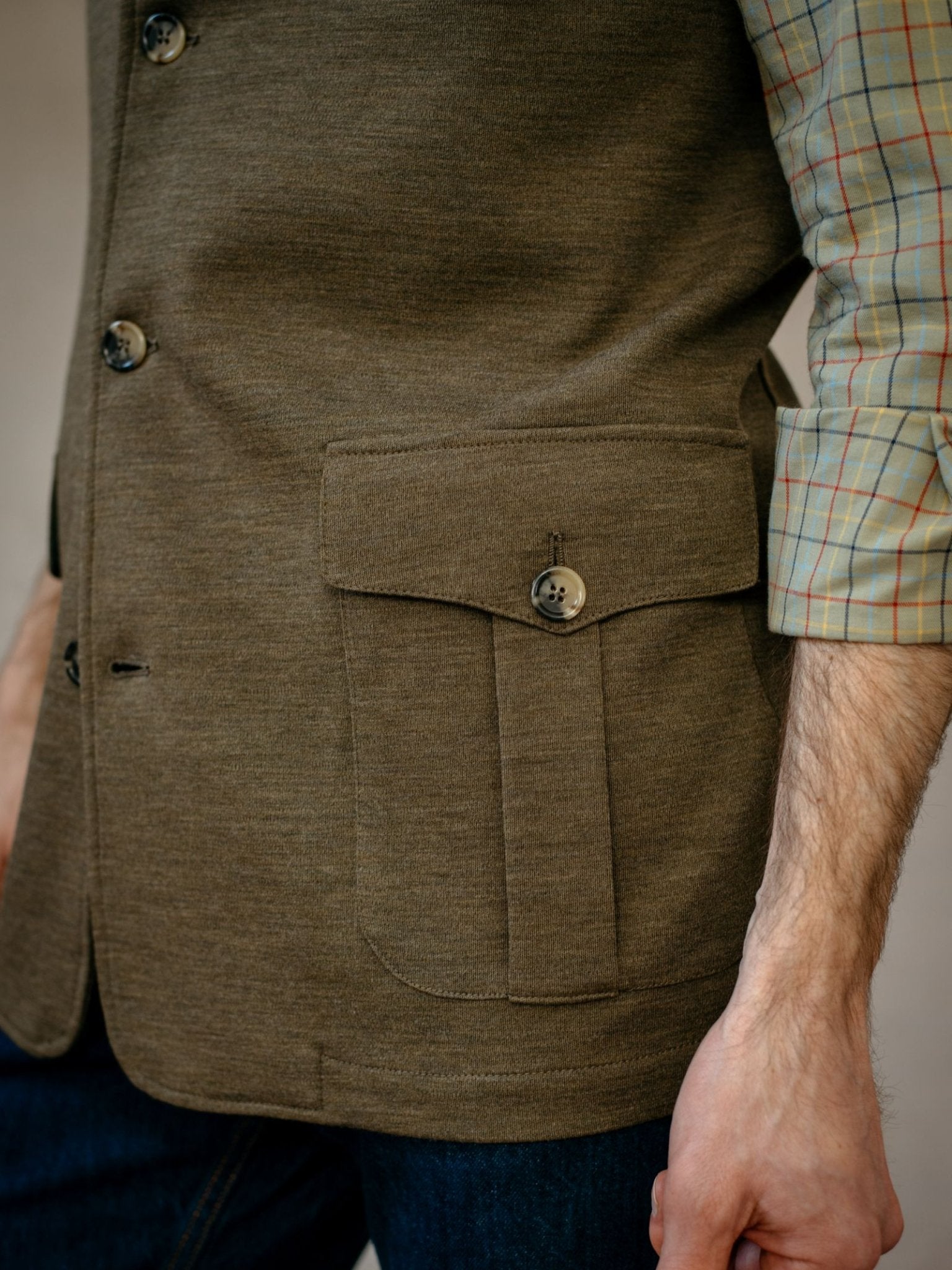Close-up of a person wearing Campbells of Beauly Knitted Nehru Gilet in olive green, 100% wool with button detail and large front pocket, styled over a beige plaid shirt with rolled sleeves. Only the torso and one arm are visible.