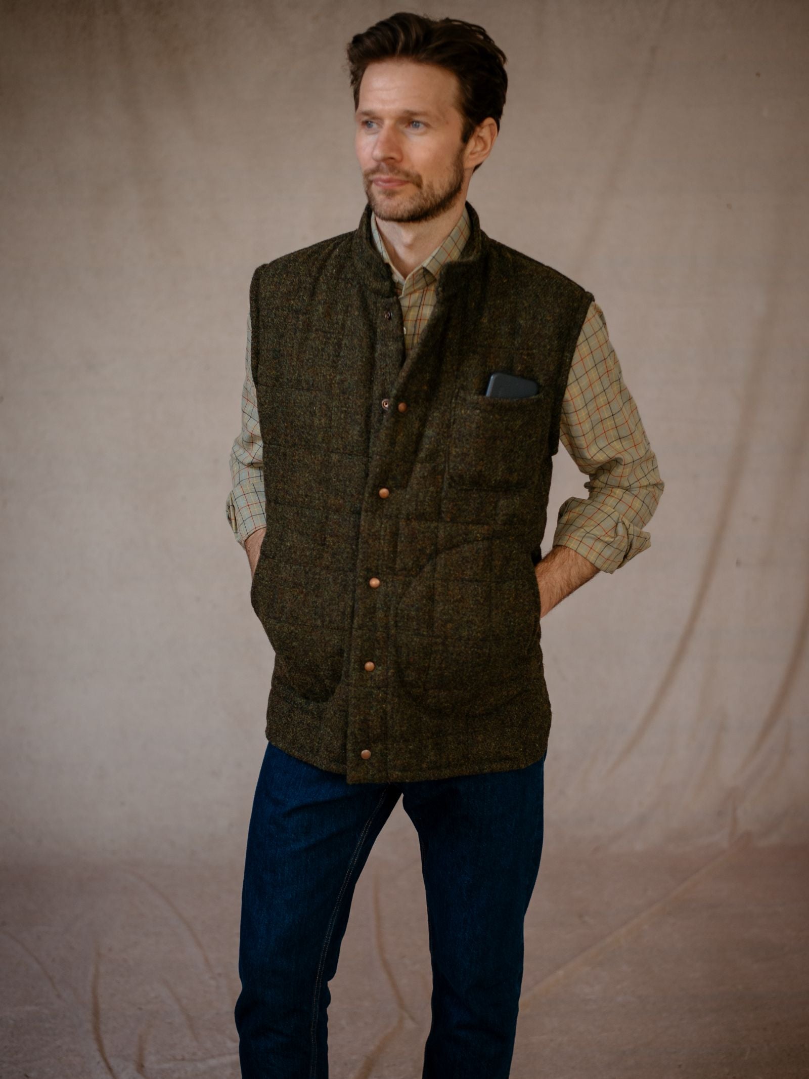 A man with short brown hair and a trimmed beard stands with hands in his pockets, wearing a plaid shirt, blue jeans, and the Campbells of Beauly Harris Tweed Quilted Gilet against a neutral, textured backdrop.