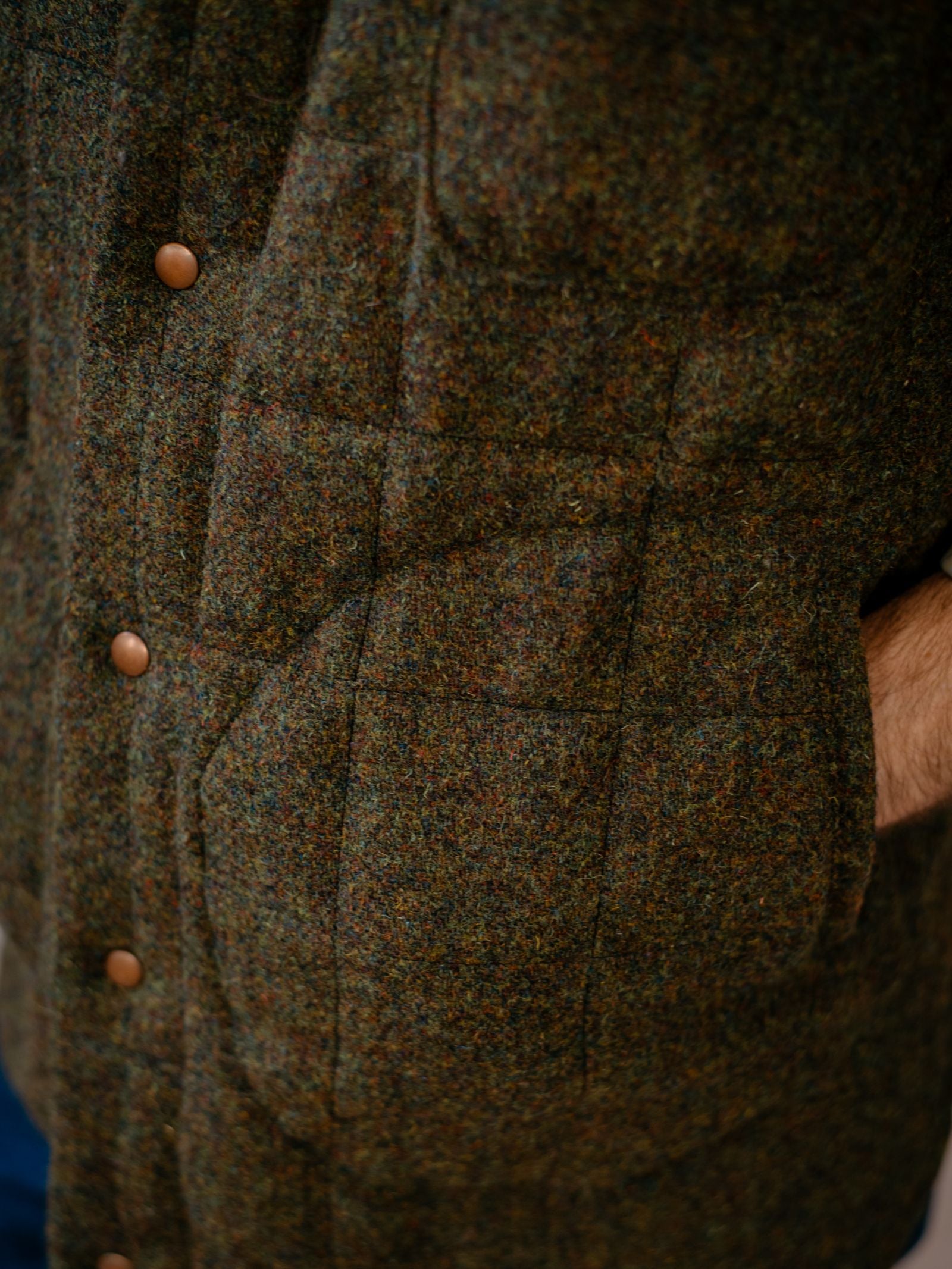 Close-up of a person wearing a Harris Tweed Quilted Gilet by Campbell's of Beauly, featuring textured brown and green fabric with brown buttons and a hand partly tucked in the side pocket.