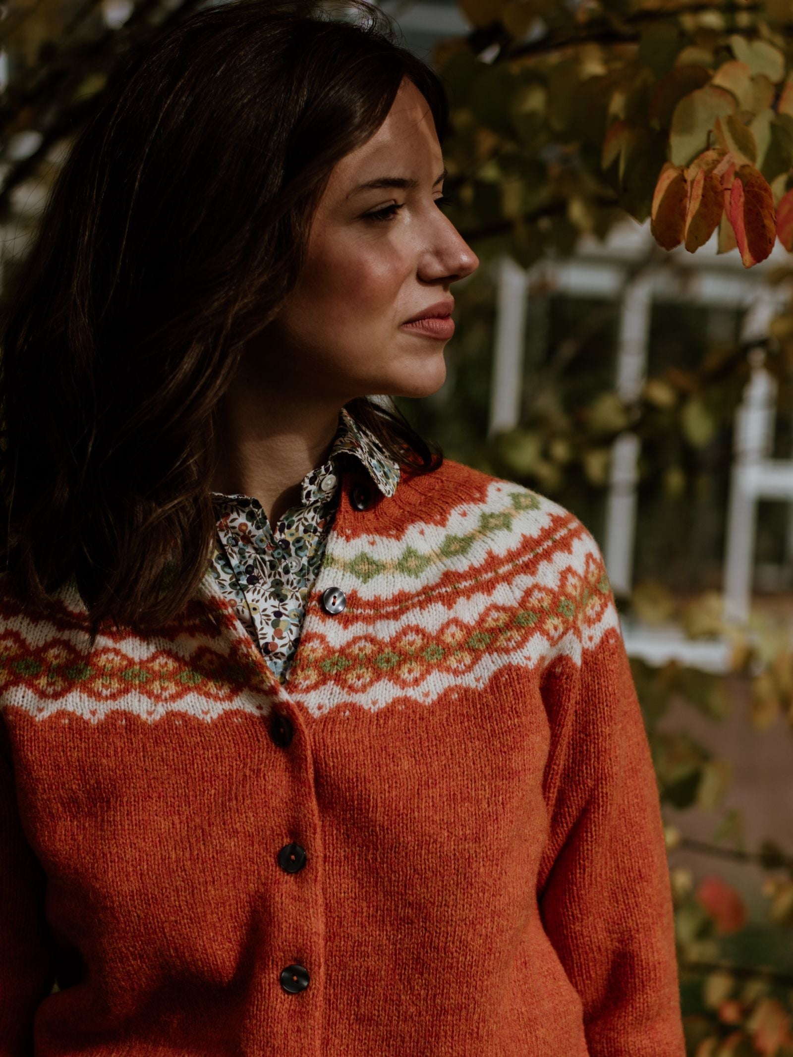 A woman with wavy brown hair stands outdoors among autumn leaves, looking to the side, wearing Campbell's of Beauly's Shetland Fairisle Cardigan over a floral shirt.