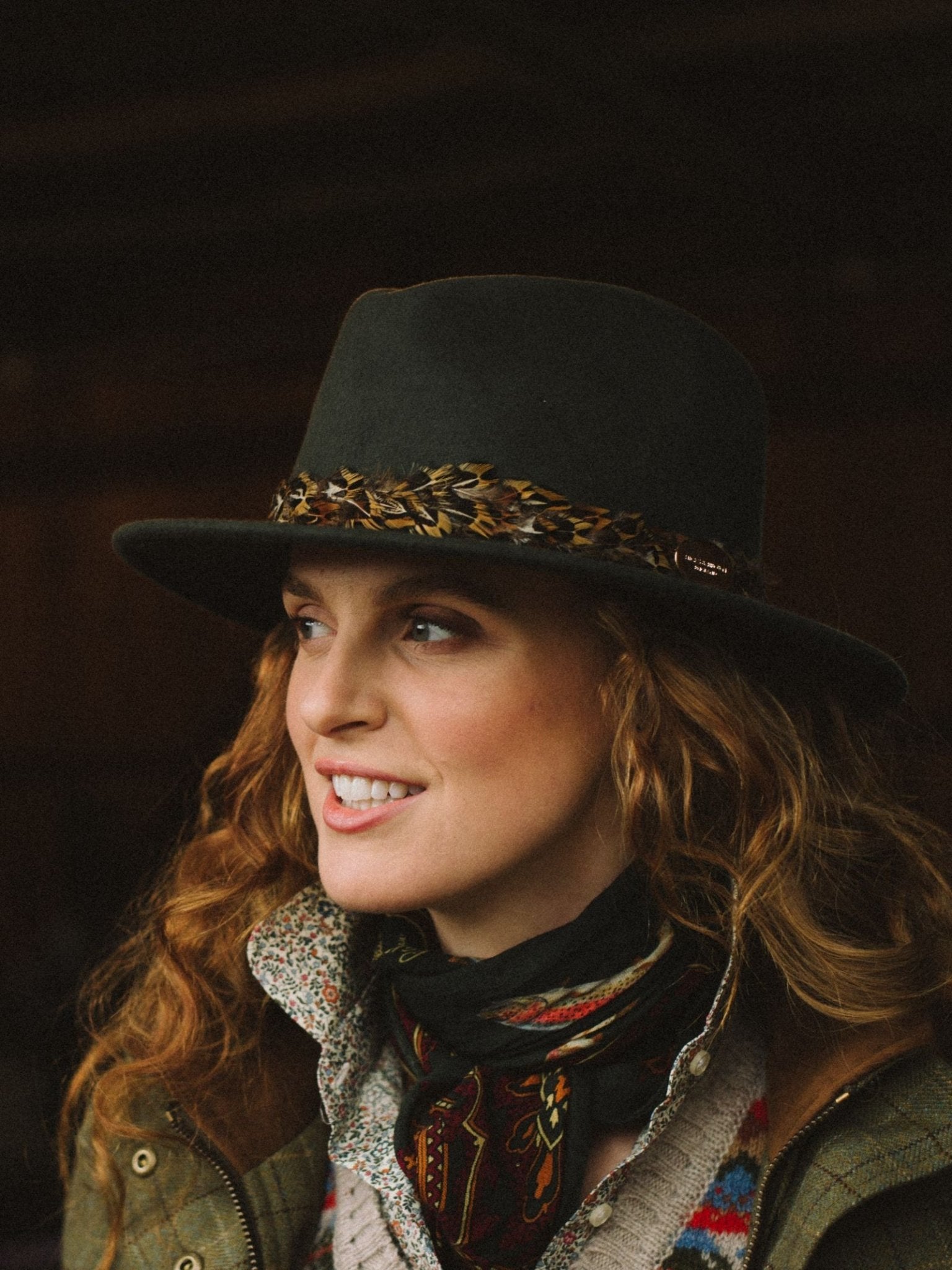 A woman with wavy red hair smiles to the side, wearing a dark wide-brimmed hat, knit sweater, green jacket, and a Campbells of Beauly Wool & Silk Scarf, standing against a dark background.