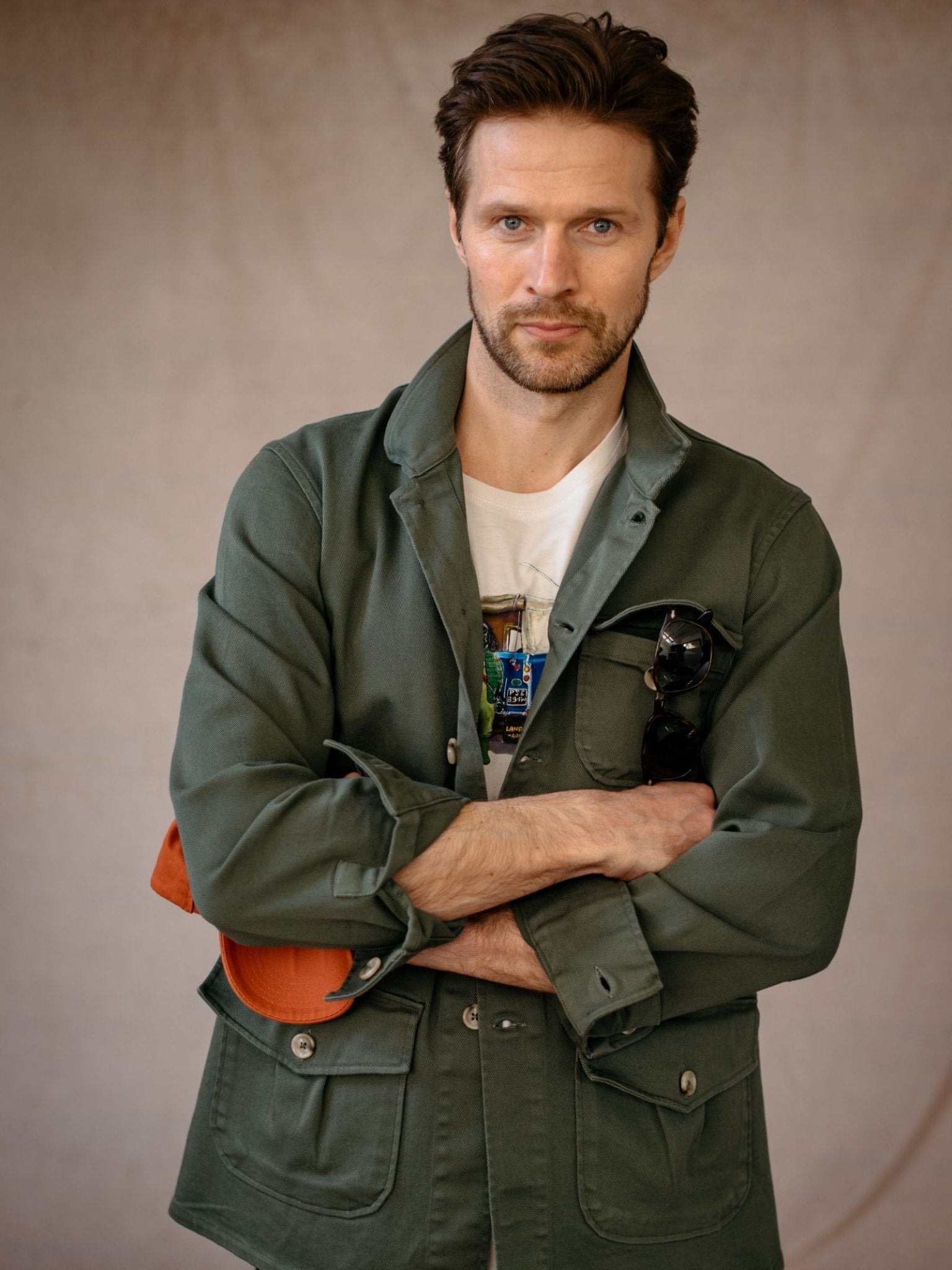 A man with light brown hair and a beard stands with arms crossed, wearing a green Campbells of Beauly Field Chore Jacket over a white t-shirt, holding sunglasses and an orange cap, looking at the camera against a plain background.
