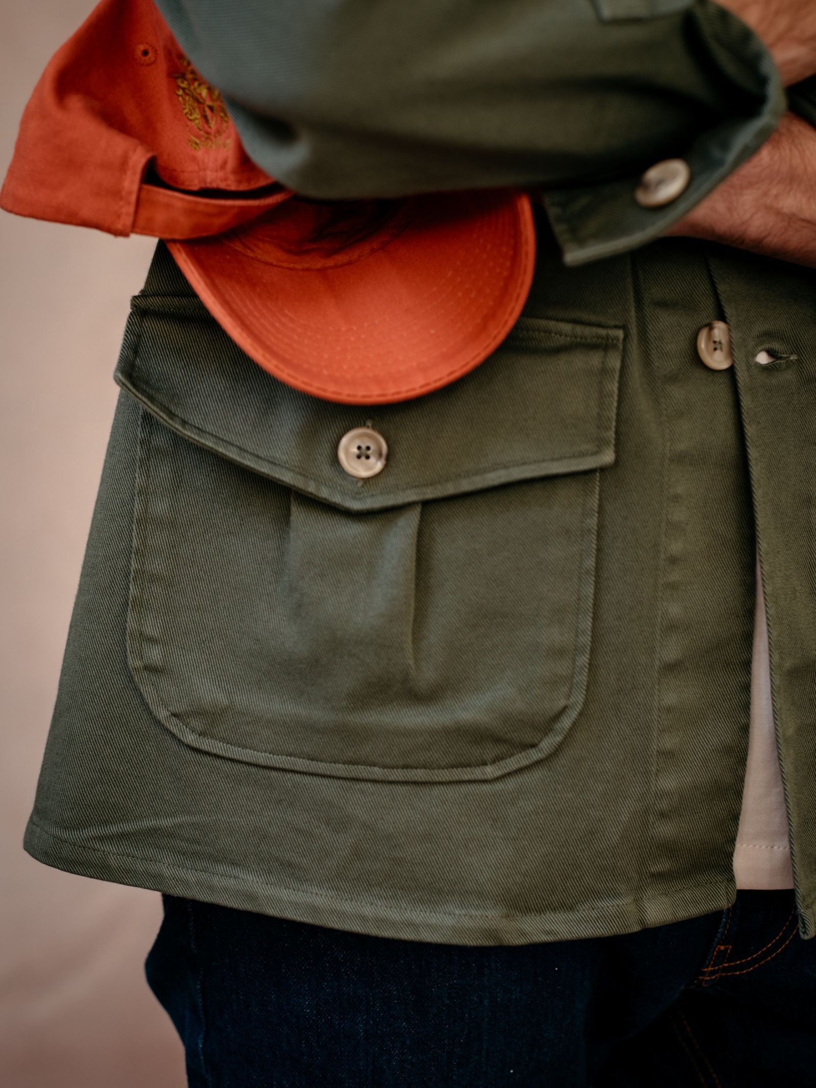 A person in a green cotton Campbells of Beauly Field Chore Jacket holds an orange cap under their arm, showing only part of their torso, arm, and the jackets utility pocket against a blurred neutral background.