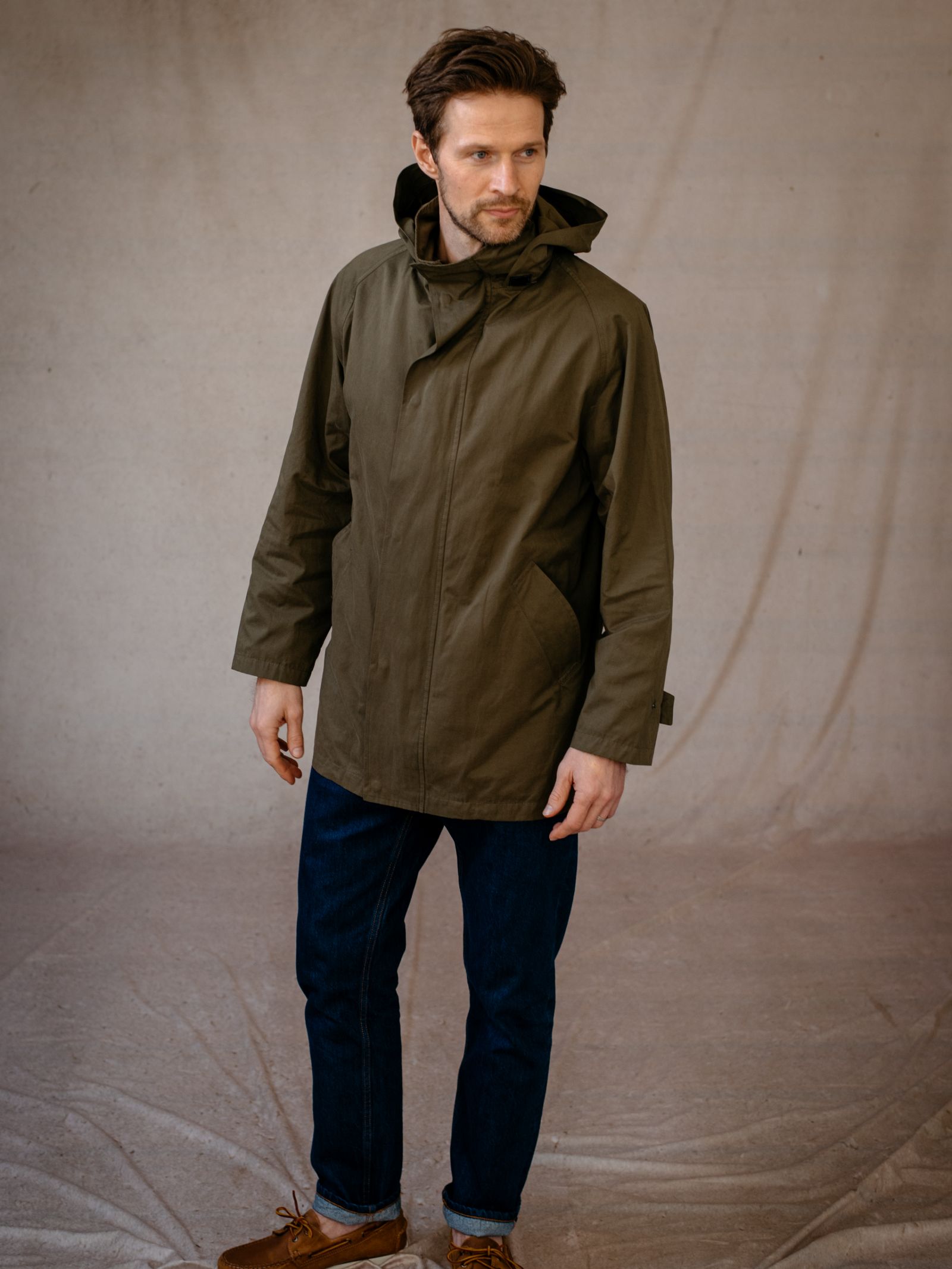 A man with short brown hair and a trimmed beard faces slightly left, wearing an olive green Campbells of Beauly Mont Coat, dark blue jeans, and brown shoes. The softly lit neutral background emphasizes the coat’s versatile, year-round style.