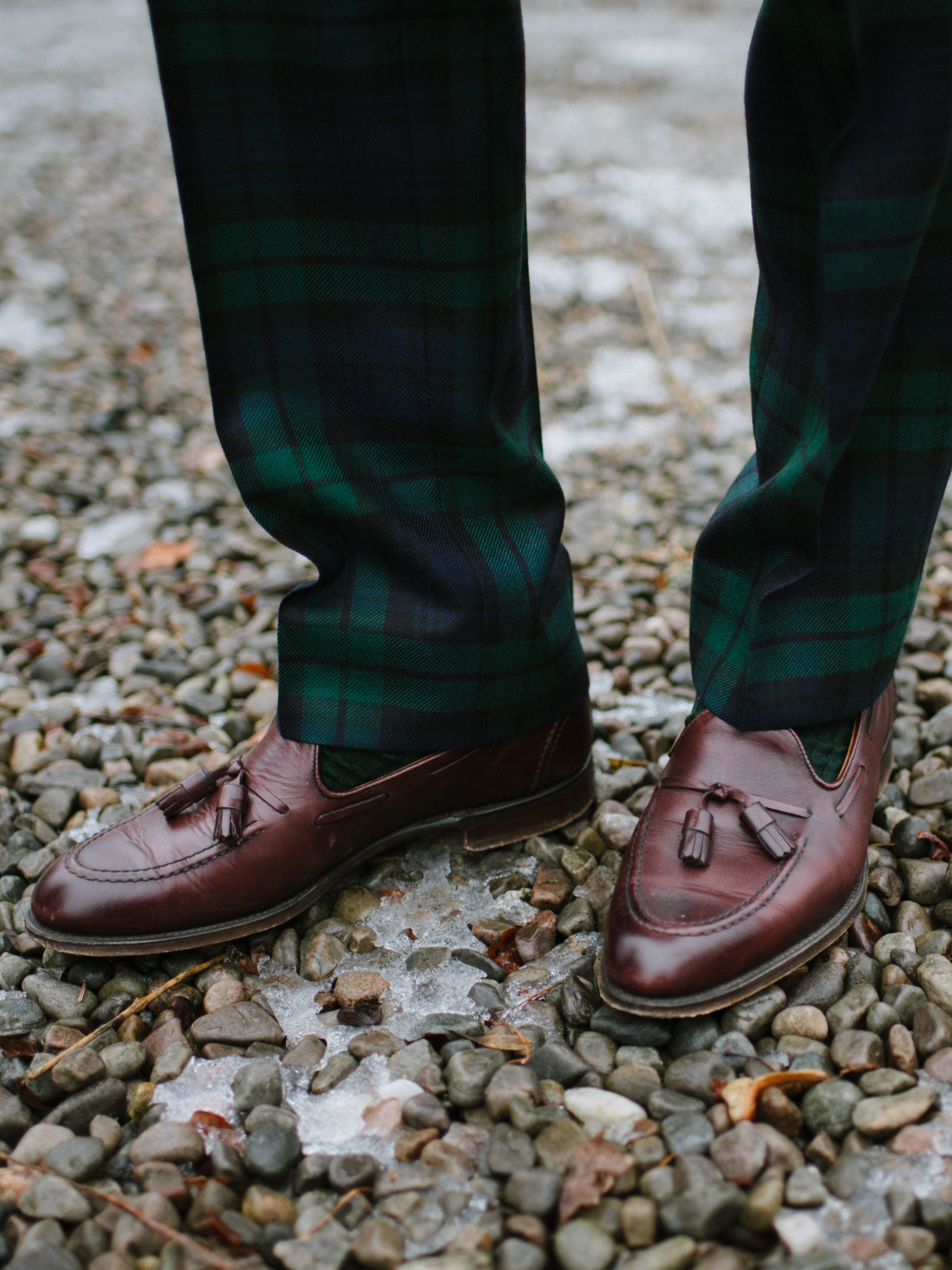 A person in Campbells of Beauly Tartan Trousers and brown leather tassel loafers stands on gravel and icy ground.
