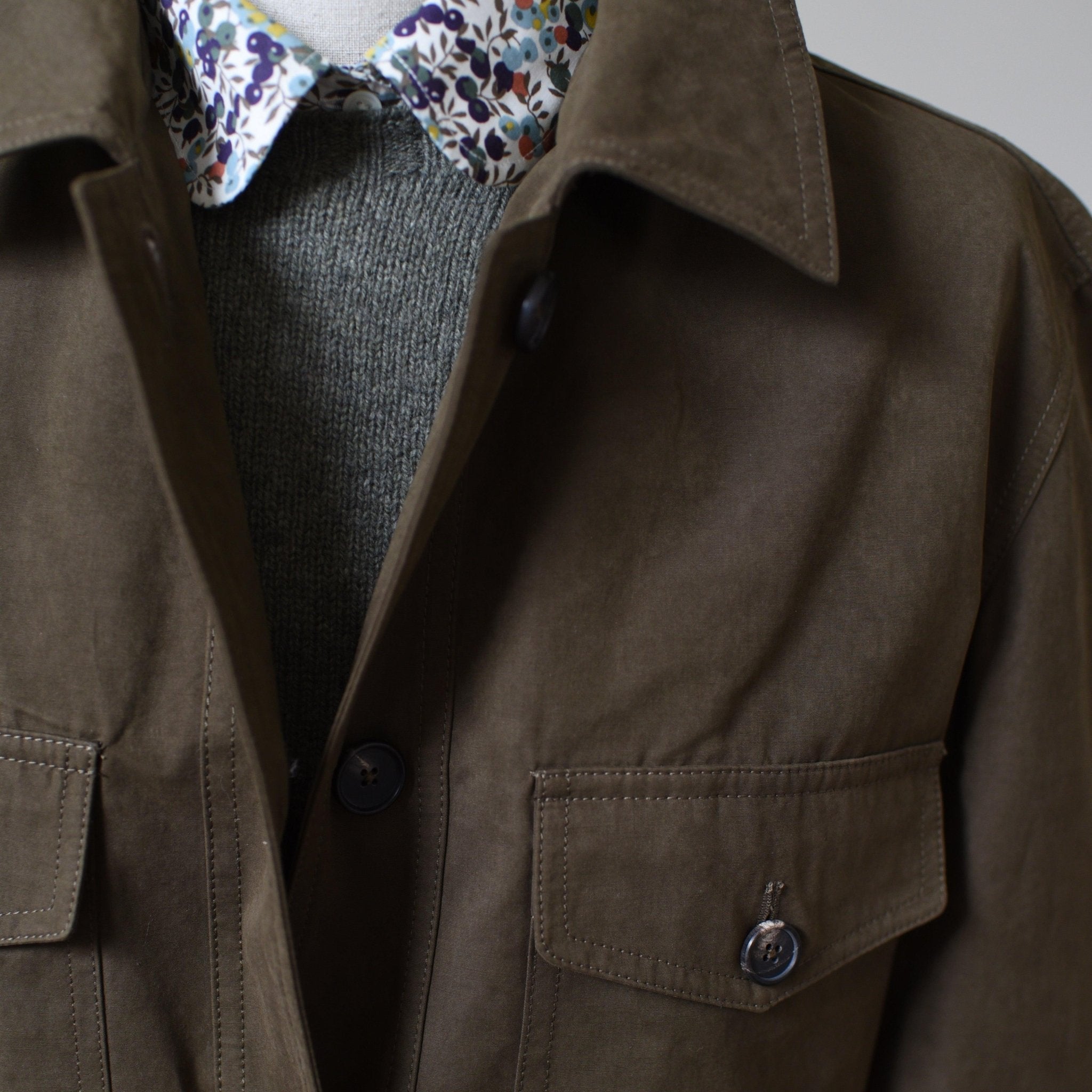 A close-up of a person wearing the Aigas Khaki Field Jacket by Campbells of Beauly, featuring buttoned pockets, layered over a grey knit sweater and a white shirt with a colorful floral pattern collar.