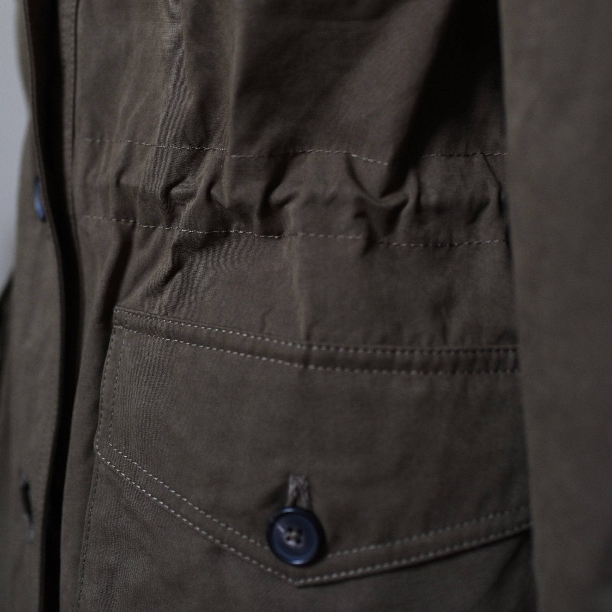 Close-up of the Aigas Khaki Field Jacket by Campbells of Beauly, featuring a buttoned pocket and stitched details. The water-repellent fabric shows a slight wrinkle, with buttons on the pocket flap and along the seam.