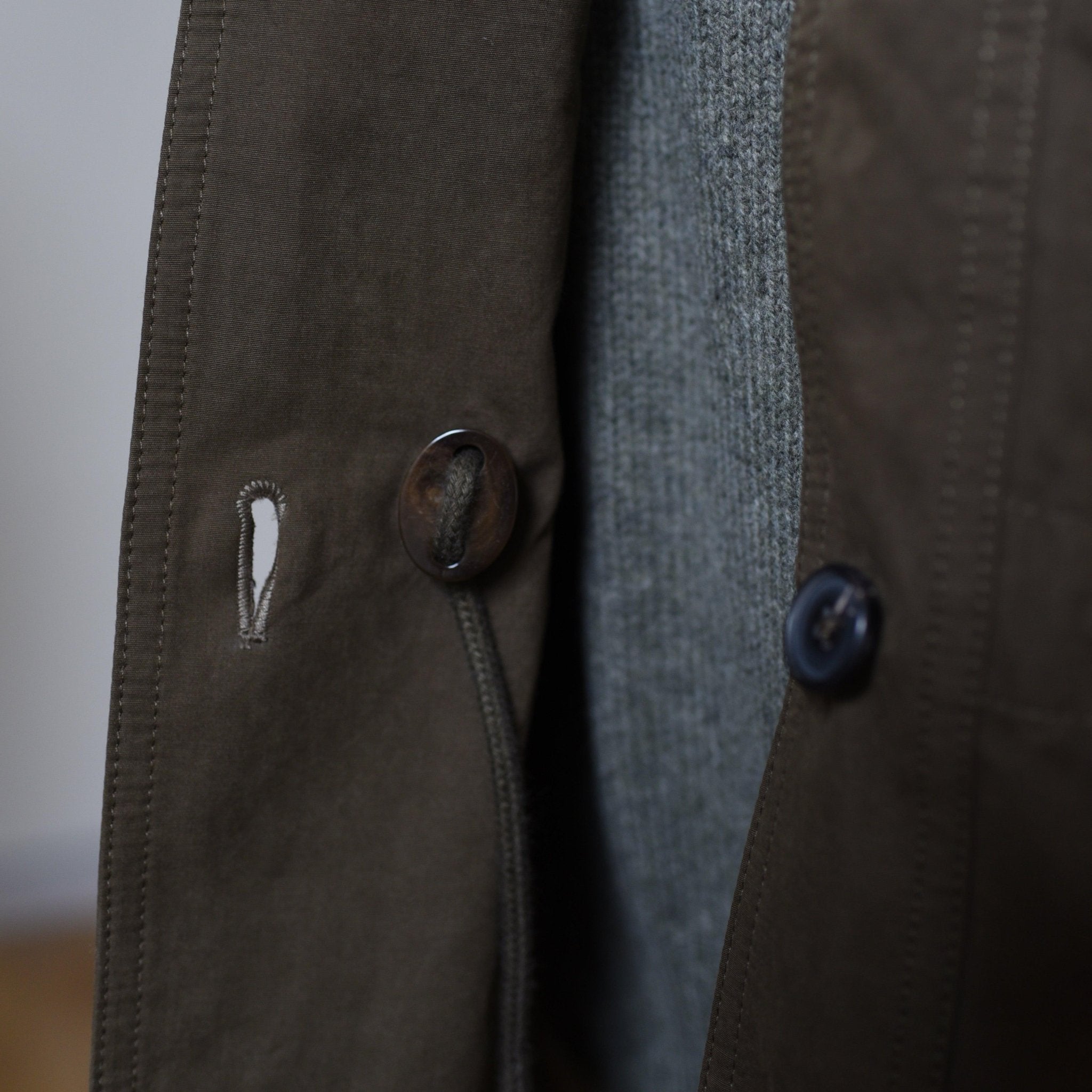 Close-up of the Aigas Khaki Field Jacket by Campbells of Beauly, showing a large dark button, detailed buttonhole, and part of a gray textured sweater beneath.