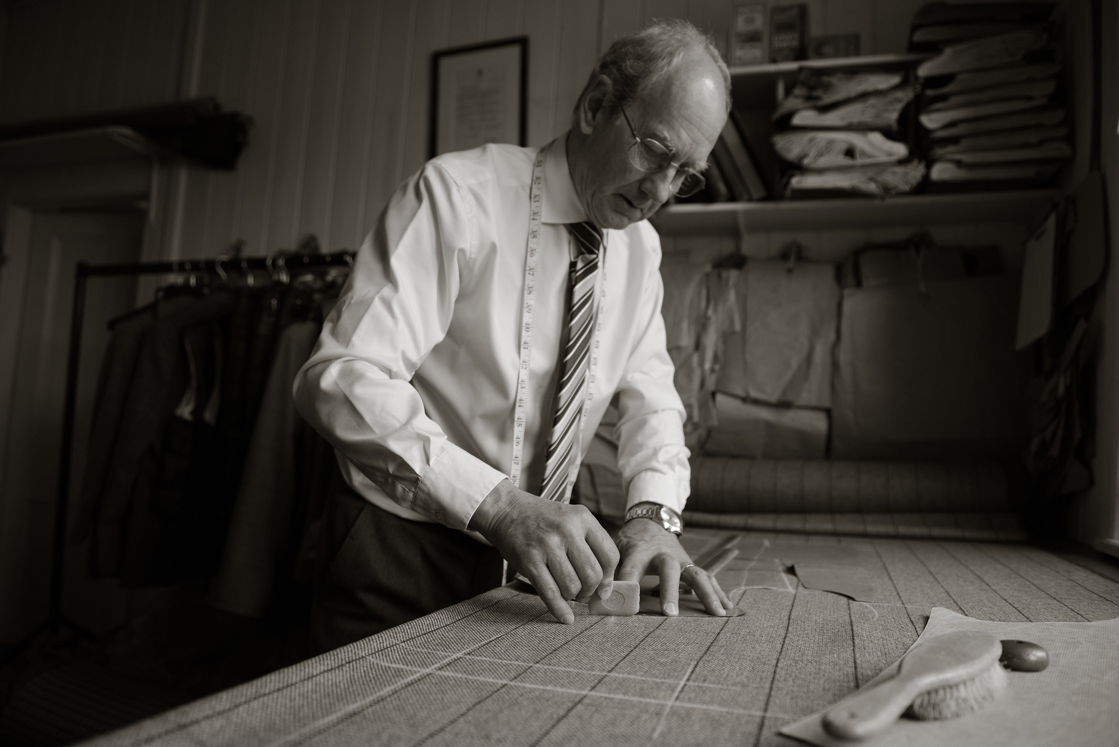 An older man in a dress shirt and tie carefully marks fabric with chalk on a worktable in a tailor’s workshop, surrounded by hanging garments and shelves of folded cloth.