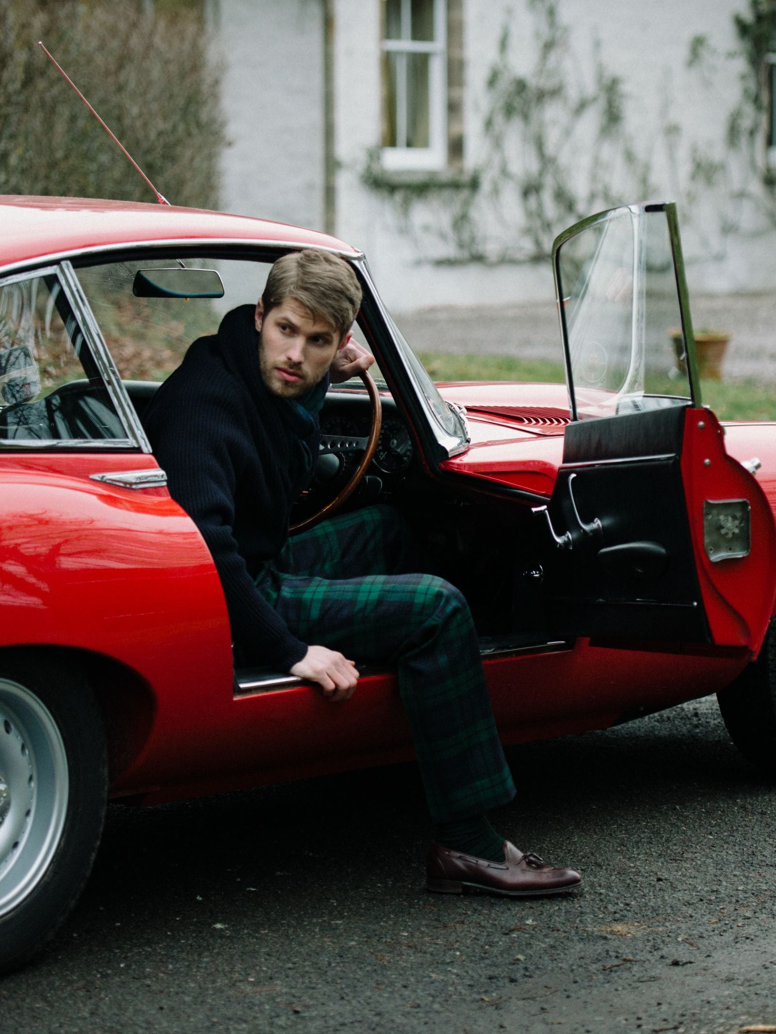 A man in a black sweater and Campbell’s of Beauly Tartan Trousers sits in the driver’s seat of a classic red car with the door open, looking left. A white building and greenery are visible in the background.