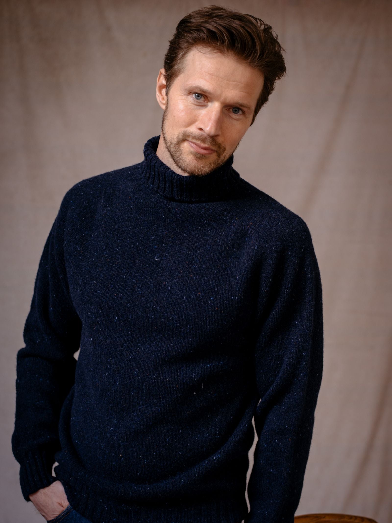 A man with short brown hair and a trimmed beard wears the Campbells of Beauly Donegal Lambswool Poloneck sweater in dark green, standing against a beige fabric backdrop with a soft, neutral expression.