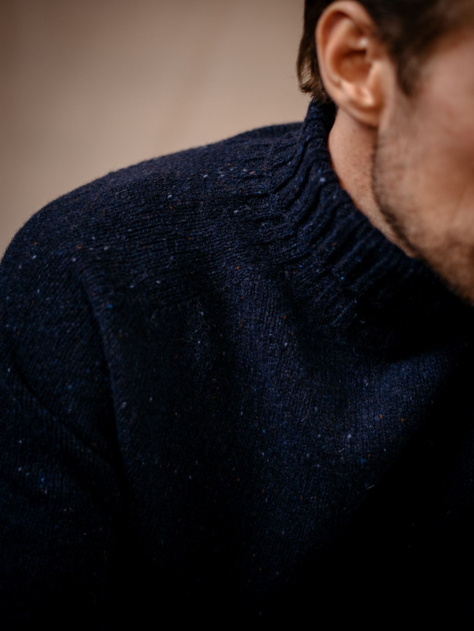 Close-up of a person wearing the Campbells of Beauly Donegal Lambswool Poloneck, a dark blue textured knit sweater with a ribbed collar, shown in focus while the background and part of their face and neck are softly blurred.