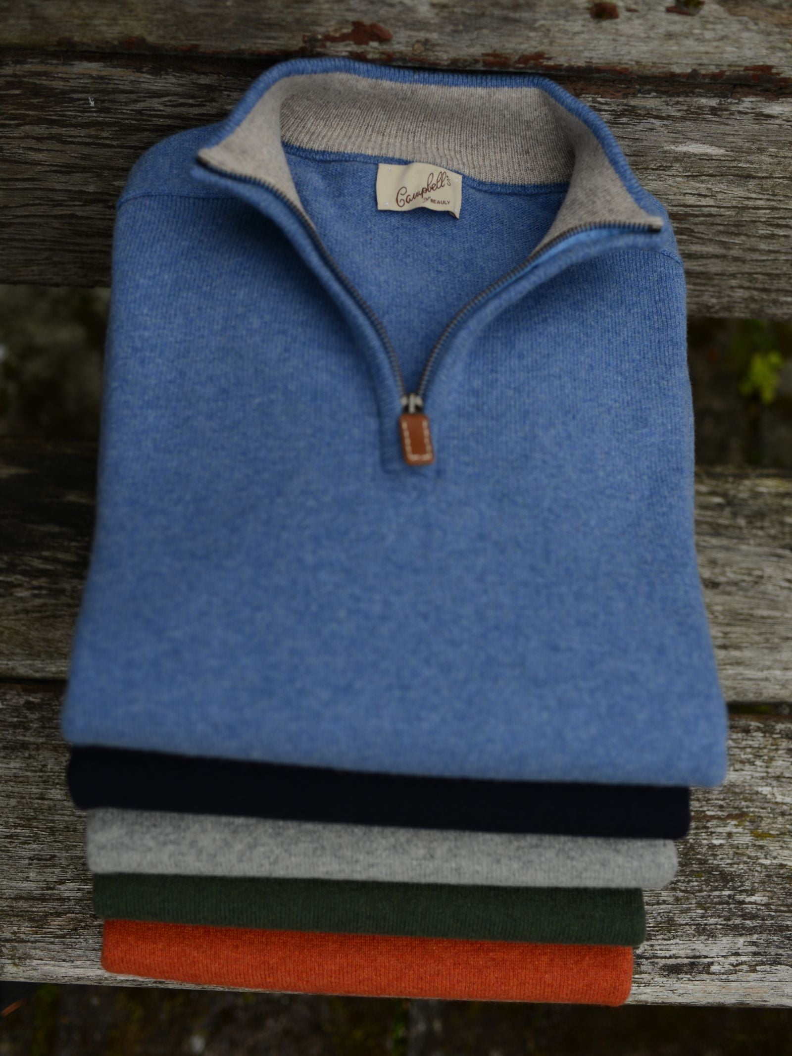 A stack of four Campbells of Beauly Lambswool Quarter Zip Jumpers in blue, navy, gray, and orange rests on a weathered bench. The top blue jumper has a light collar and a brown leather zipper pull.