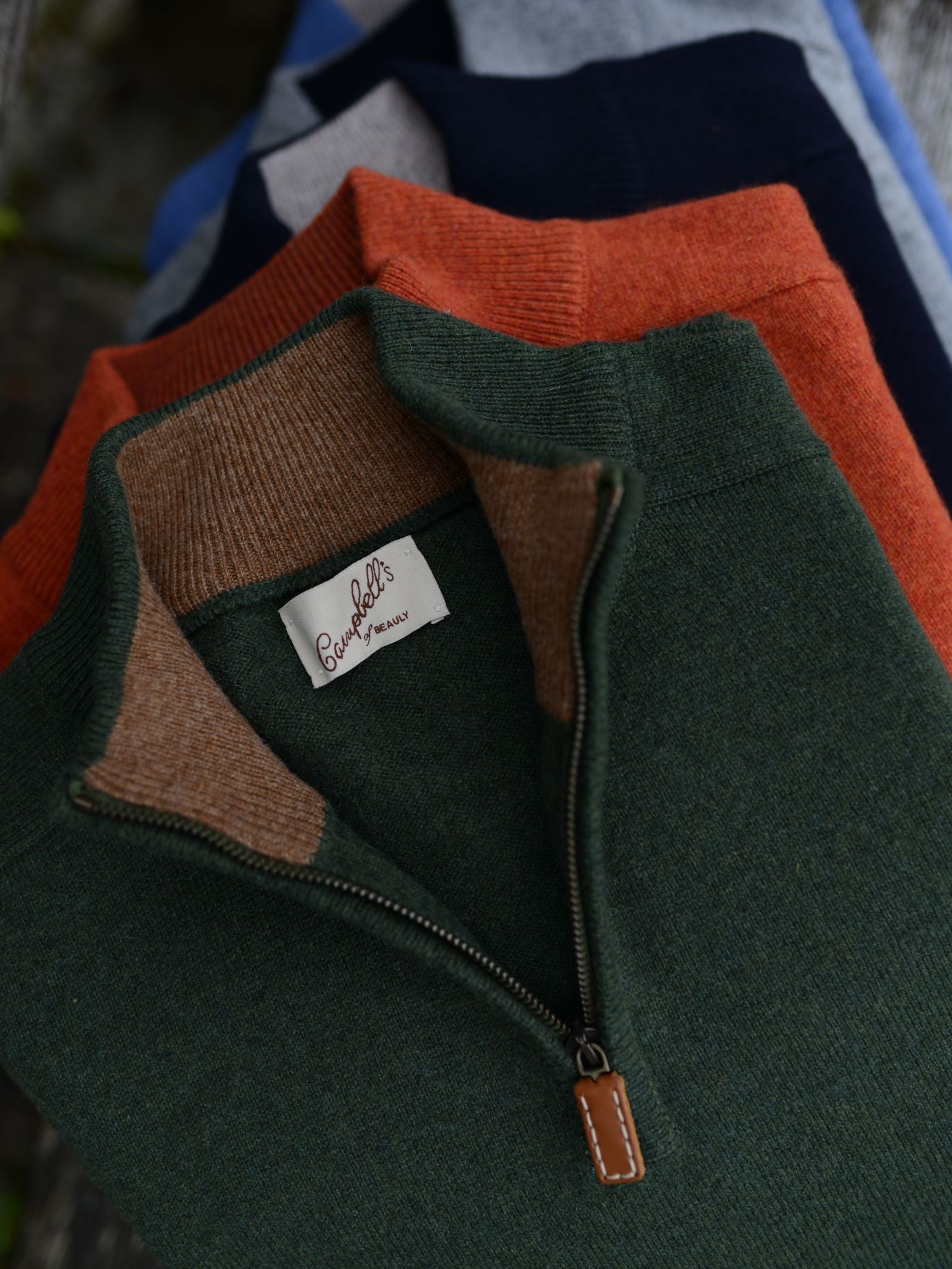 Three Lambswool Quarter Zip Jumpers by Campbells of Beauly in dark green, orange, and navy with contrasting collars are stacked. The top green jumper shows a “Baruffa” label and brown zipper pull. Inspired by the Scottish Borders.