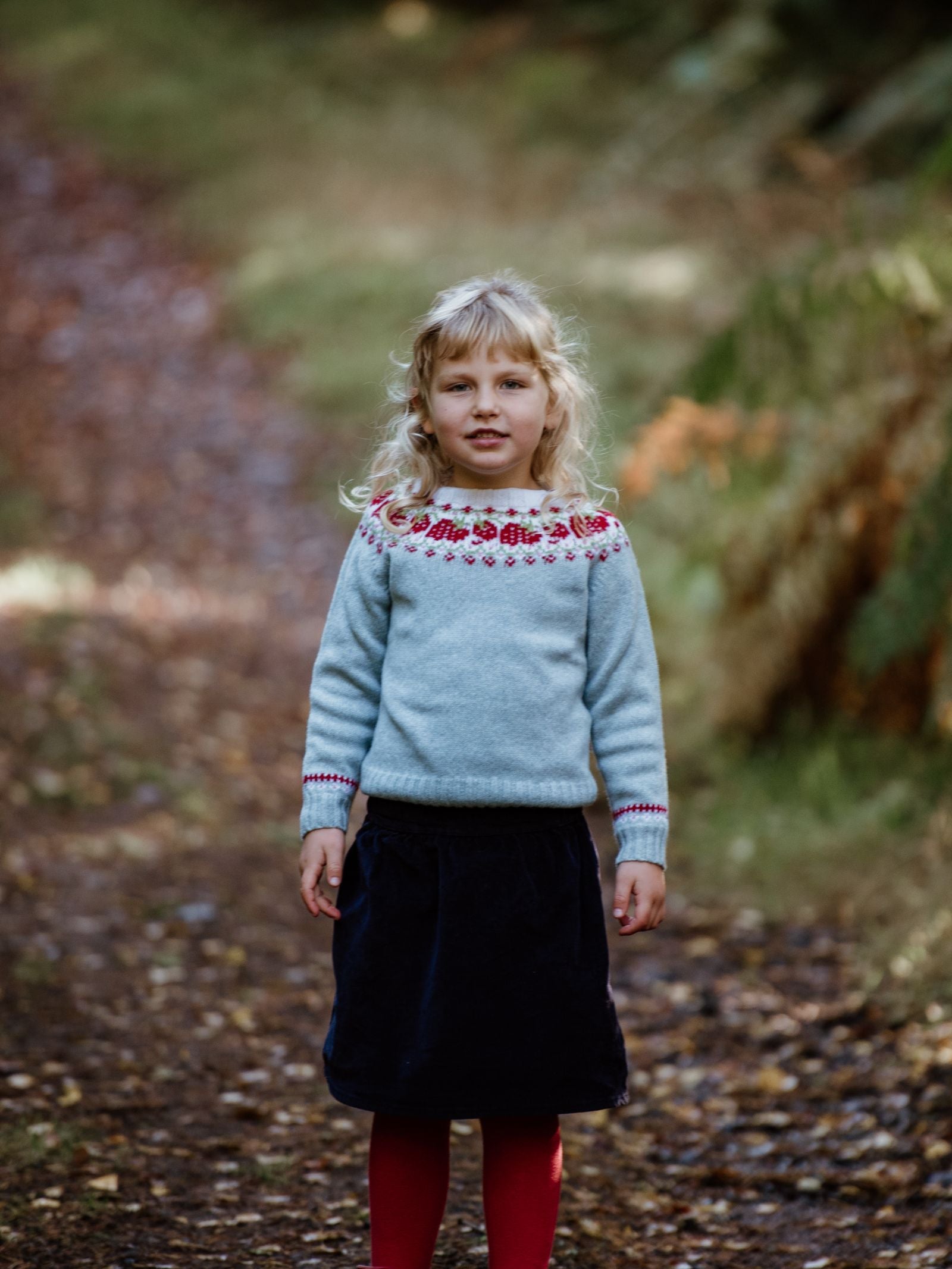 A young girl with blond hair stands on a woodland path, wearing the Campbells of Beauly Mini Strawberry Yoke Jumper, paired with a dark skirt and red tights. The background features blurred trees and fallen leaves.
