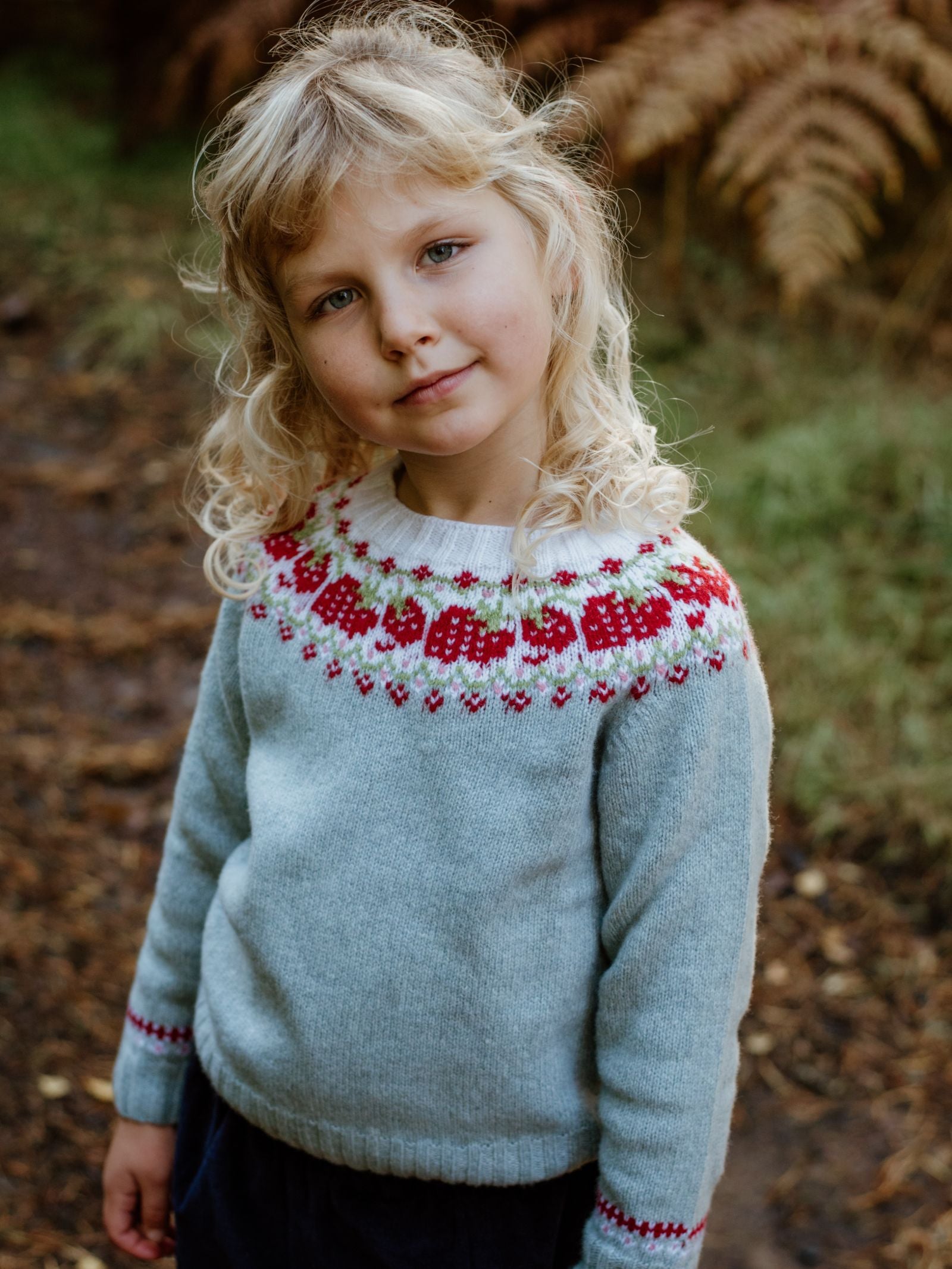 A young child with wavy blonde hair stands outdoors in a Campbells of Beauly Mini Strawberry Yoke Jumper, featuring a light blue base and red, white, and pink Fairisle yoke. Ferns and greenery blur in the background.