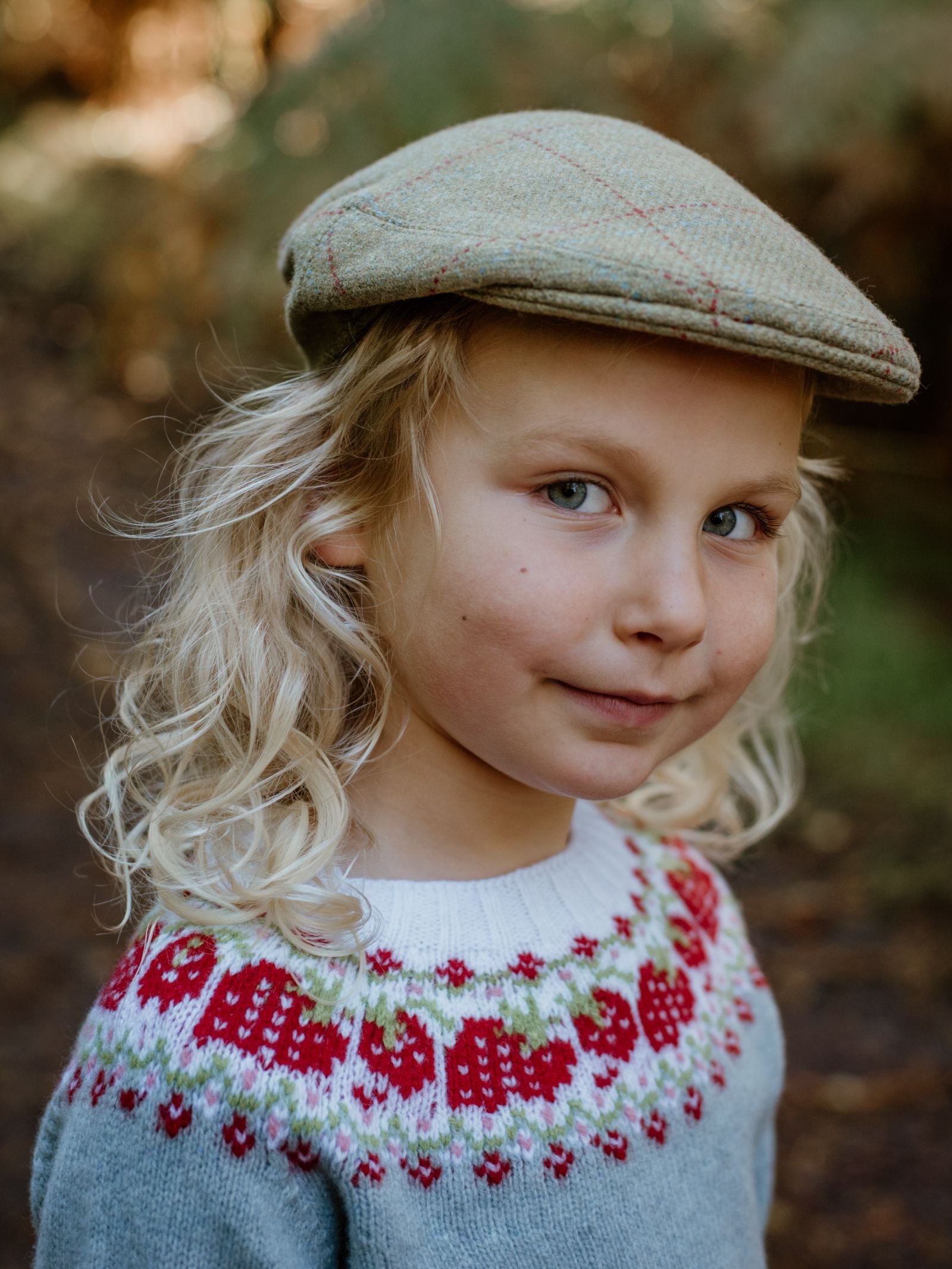 A young child with long blond hair smiles softly while standing outdoors on a wooded path, wearing the Mini Tweed Cap by Campbells of Beauly and a light blue sweater with red and white accents—ideal for little adventurers.