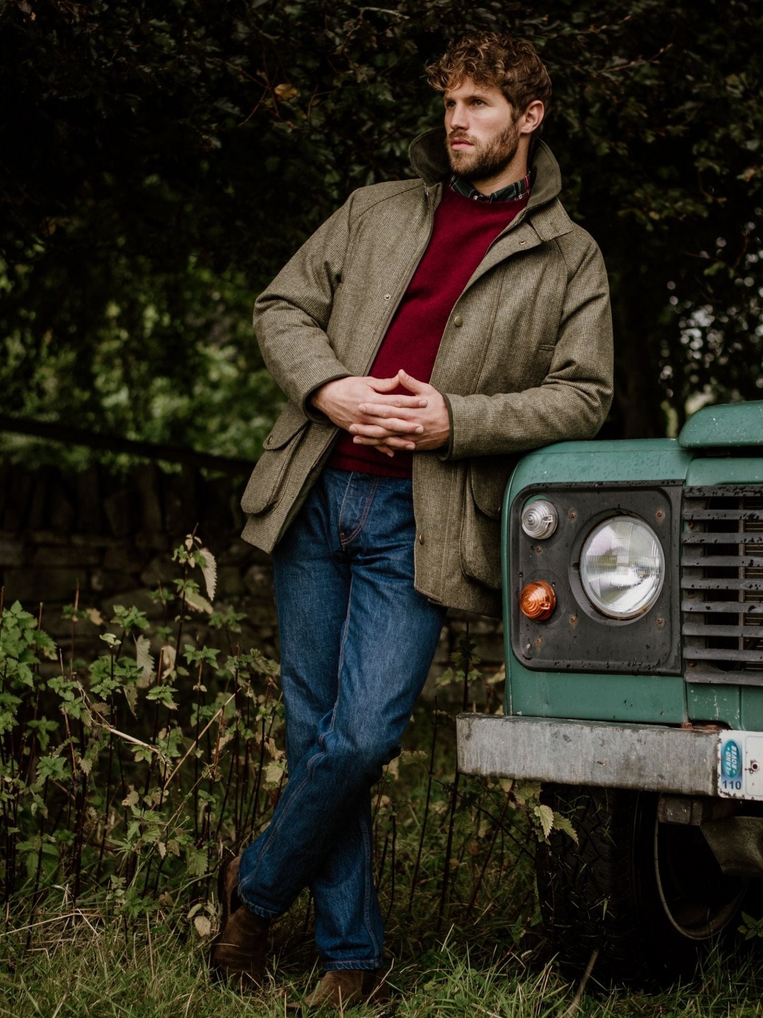 A man in a green Campbells of Beauly Tweed Field Coat with an Alcantara collar, red sweater, and blue jeans leans against an old green vehicle outdoors, surrounded by trees and lush greenery.