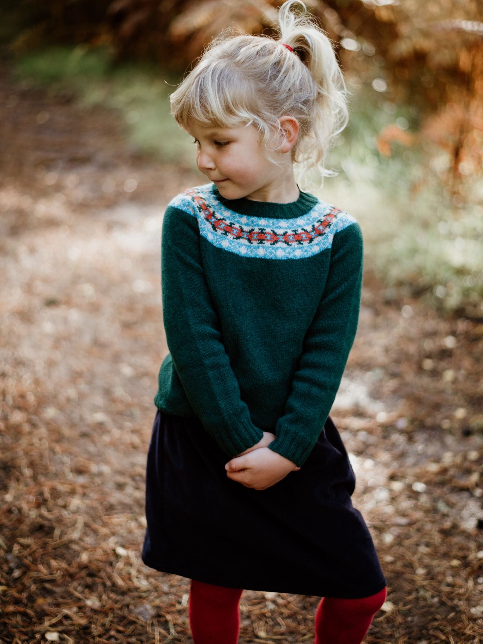 A young girl with blond hair in a ponytail stands on a leafy path, smiling softly as she wears the Campbells of Beauly Mini Fairisle Crew Jumper, styled with a navy skirt and red tights.