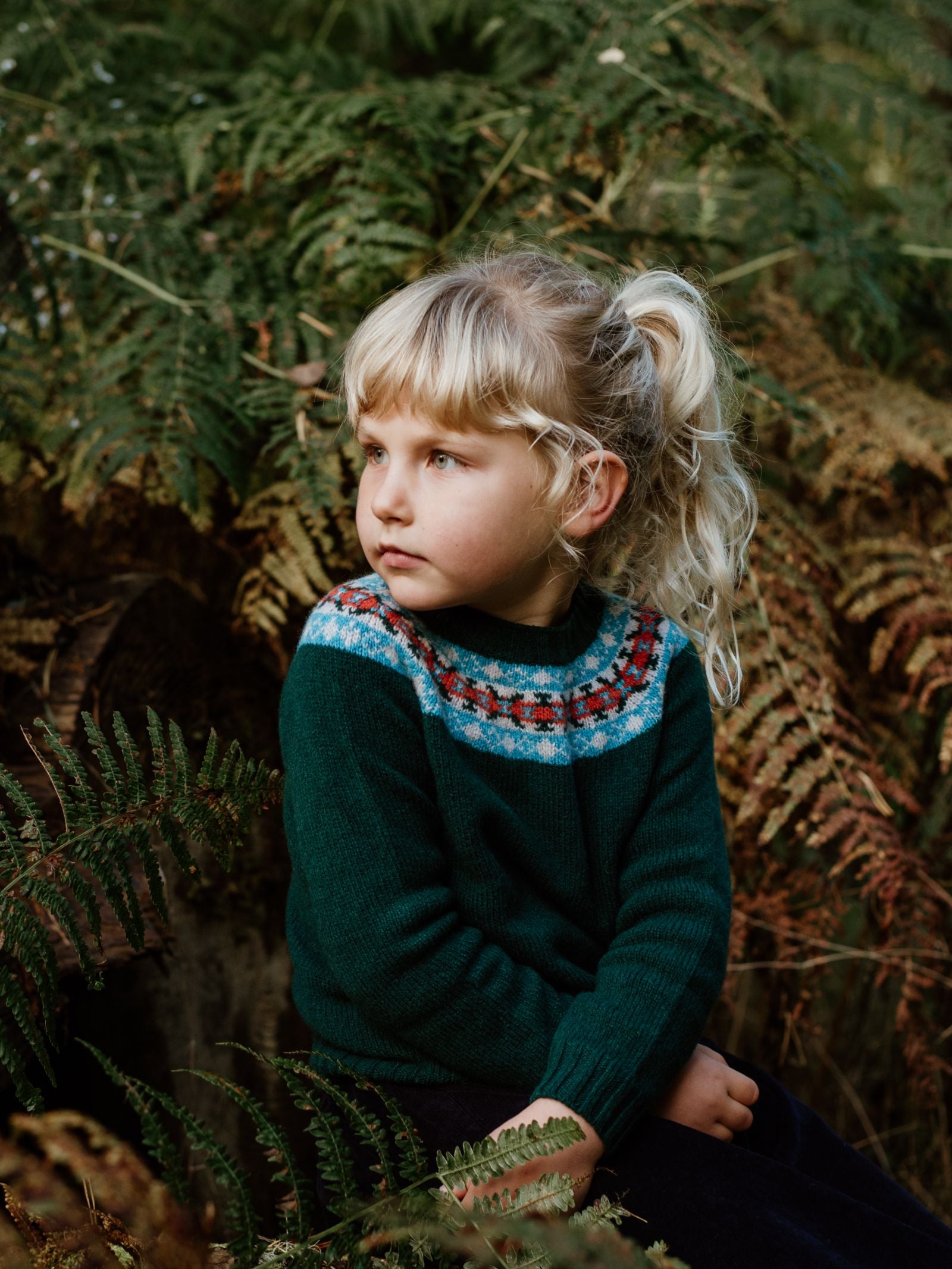 A young child with blonde hair wears a dark green Campbells of Beauly Mini Fairisle Crew Jumper with a colorful pattern, sitting among green ferns and gazing thoughtfully to the side in an outdoor setting.
