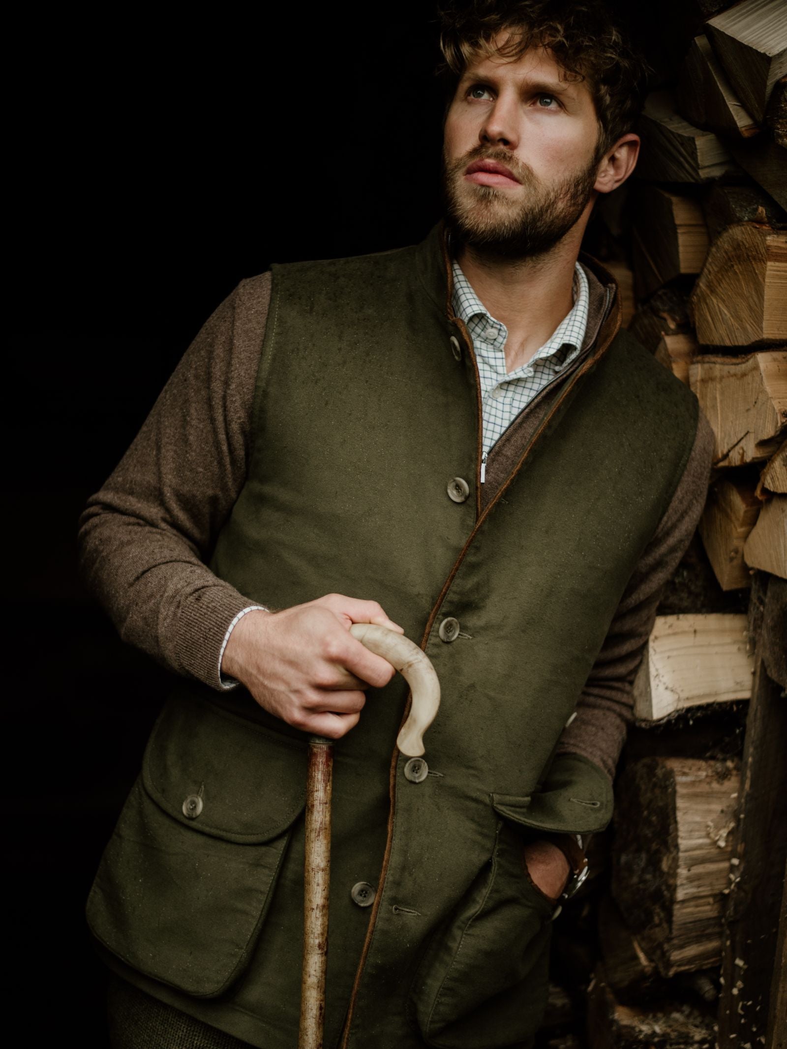 A man with curly hair and a beard, wearing the Campbells of Beauly Field Nehru Gilet in green over a brown sweater, holds a wooden walking stick and leans on firewood, gazing thoughtfully into the distance.