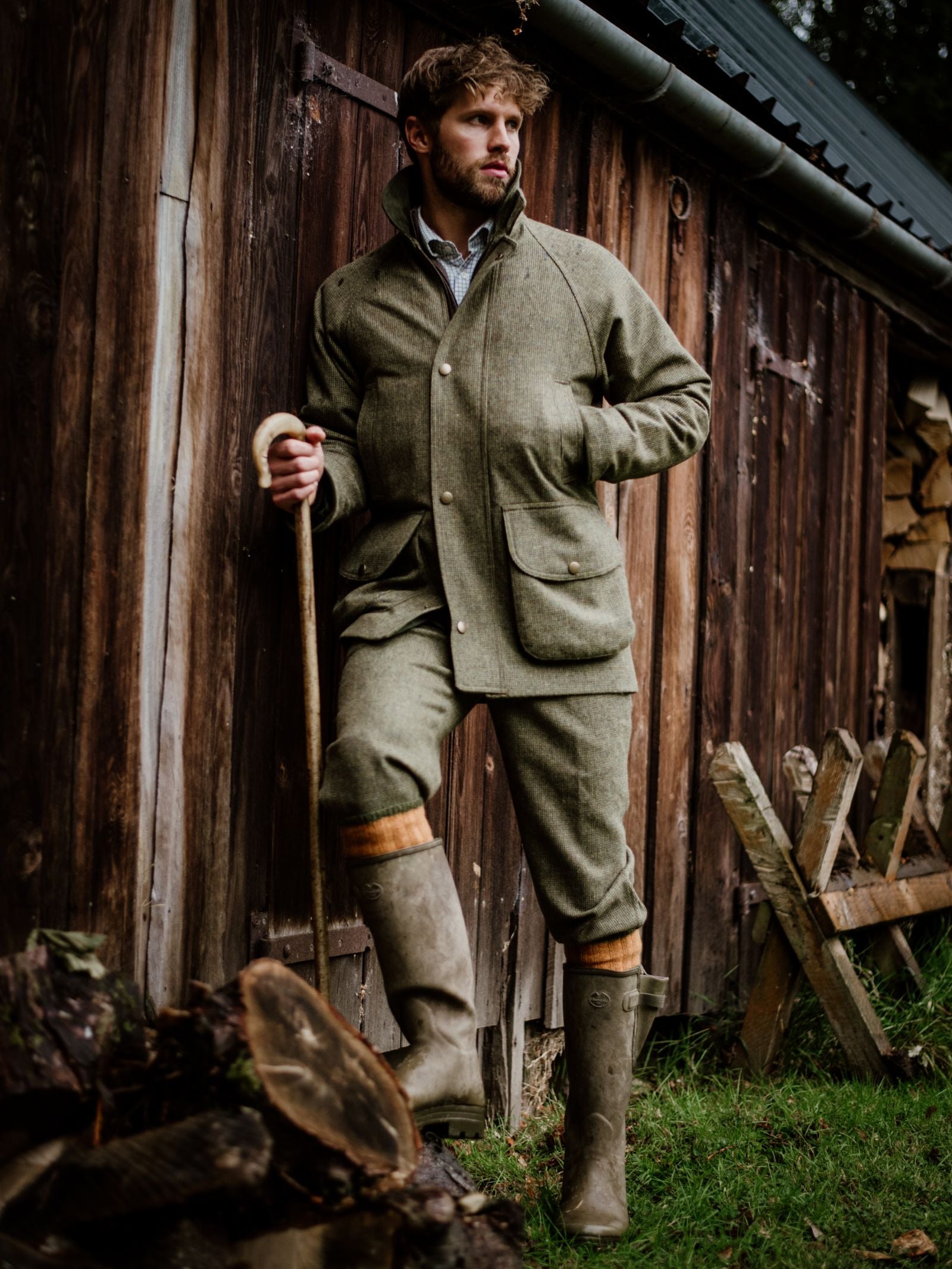 A man in countryside attire wears a Campbells of Beauly Tweed Field Coat with an Alcantara collar, matching trousers, and boots. He stands by a rustic wooden shed with a walking stick; logs and a fence are visible in the grassy yard.