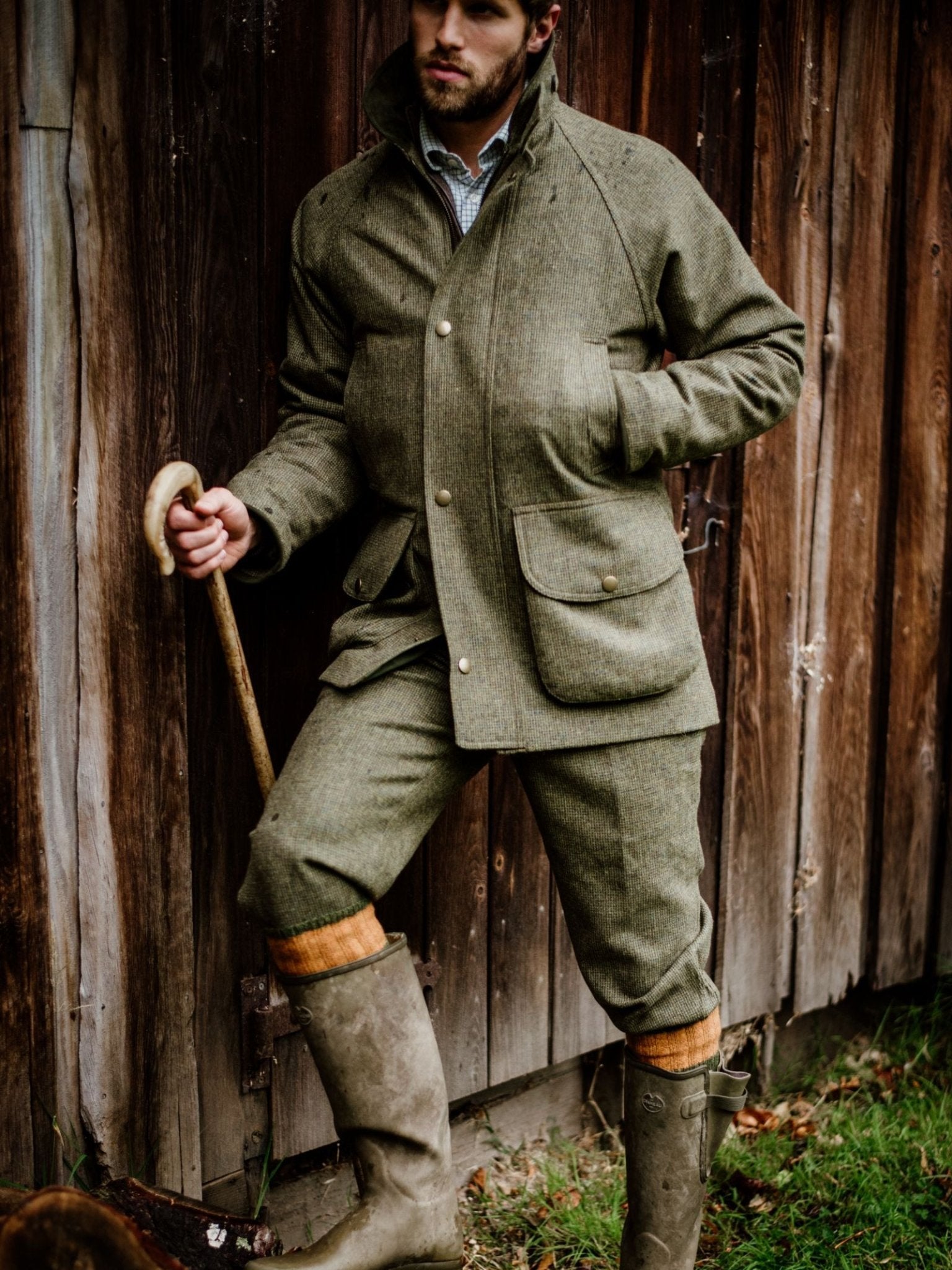 A person in Campbells of Beauly Tweed Breeks, a matching green Scottish-woven jacket, yellow socks, and rubber boots stands by a wooden wall with a cane and one hand in their pocket.